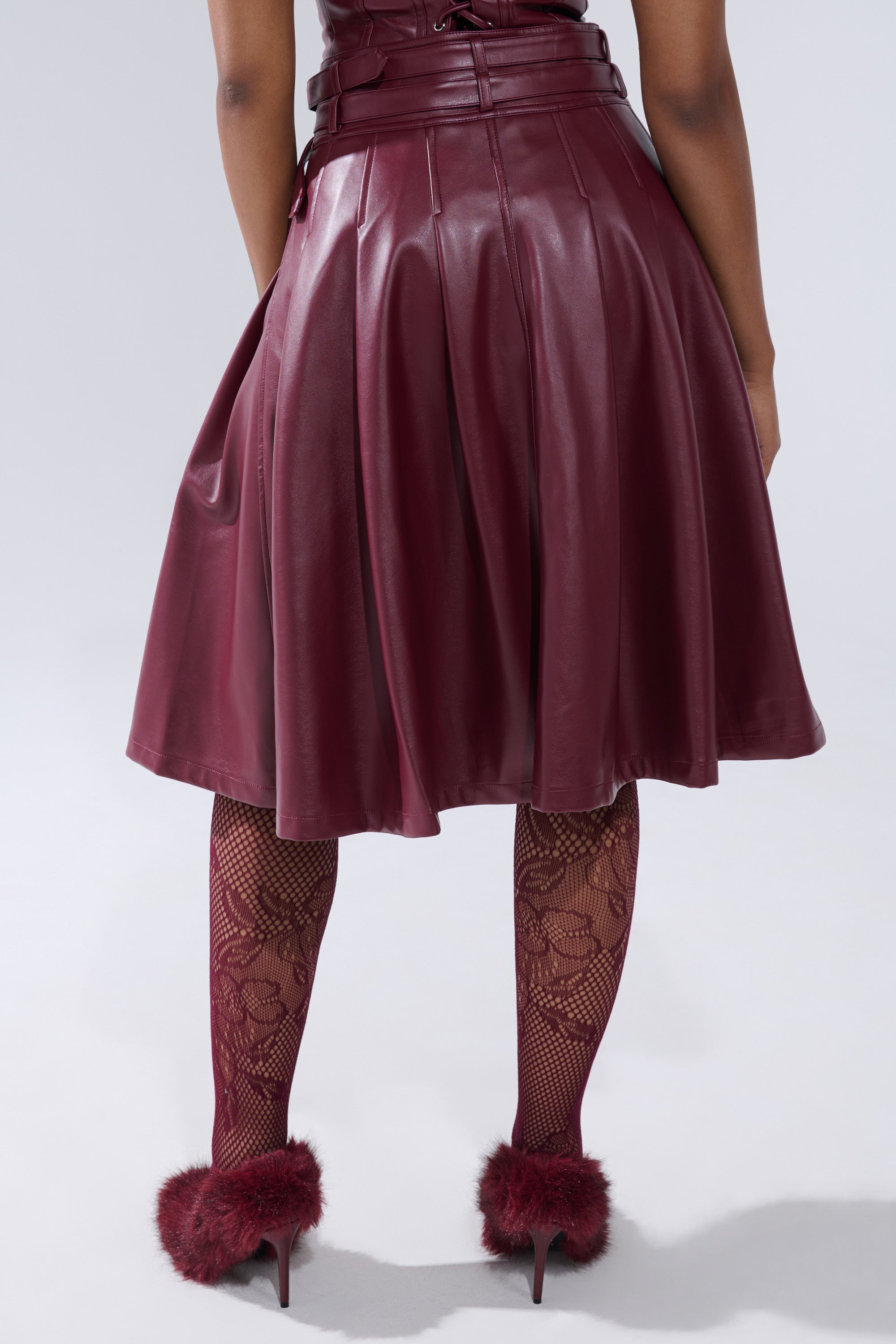 A person faces away, wearing the FEELING THE ENERGY MIDI SKIRT in burgundy faux leather, paired with patterned burgundy tights and matching high heels with fluffy accents. The background is plain light gray.