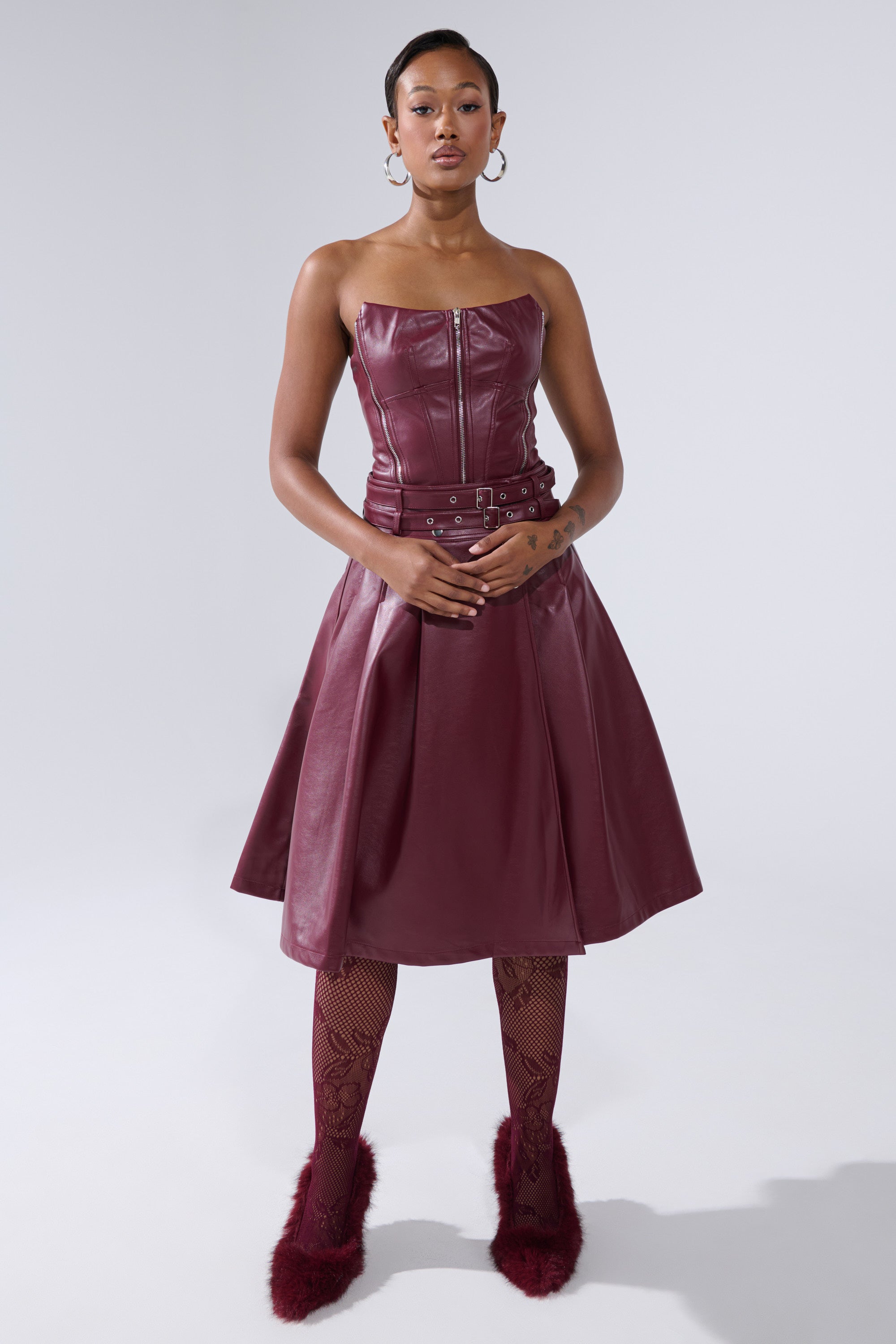 A woman poses against a plain background wearing the FEELING THE ENERGY MIDI SKIRT, featuring a strapless burgundy faux leather flared design, paired with textured tights, hoop earrings, and fluffy burgundy shoes. Her hands are gracefully clasped.