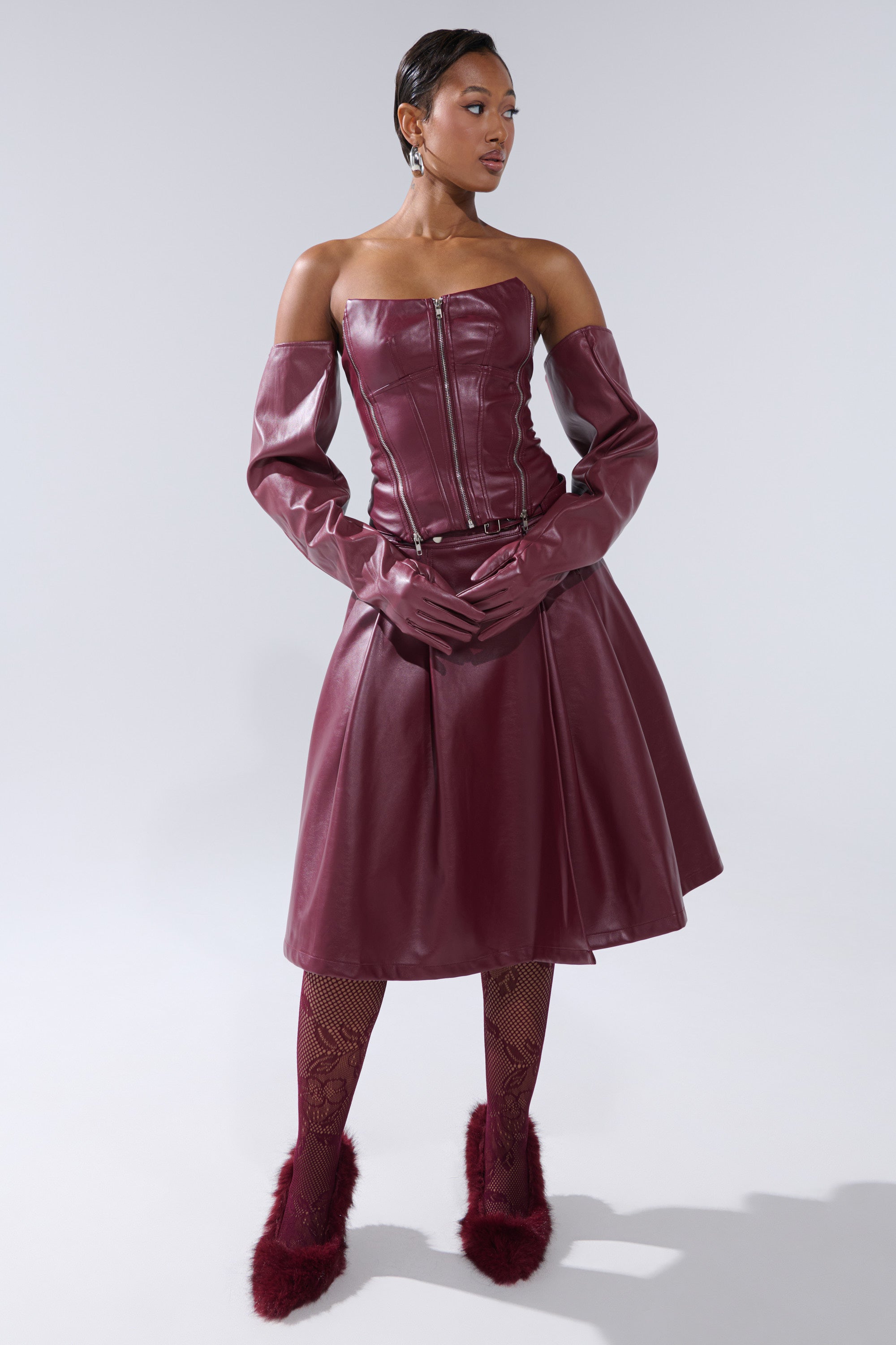 A woman poses against a plain light gray background in the FEELING THE ENERGY MIDI SKIRT, styled with a burgundy off-shoulder leather dress, long gloves, fishnet tights, and fuzzy burgundy heels.