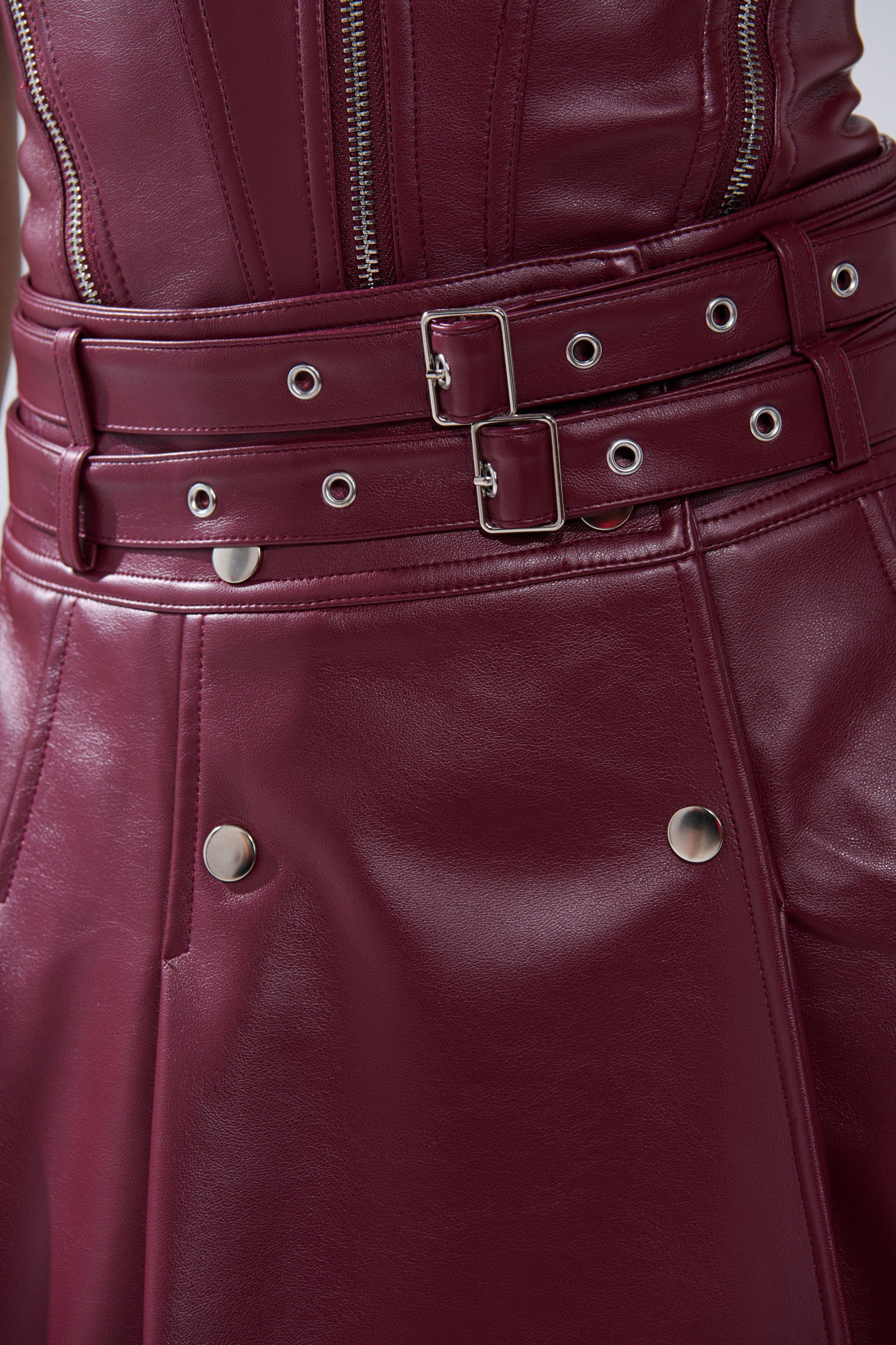 Close-up of the FEELING THE ENERGY MIDI SKIRT in burgundy faux leather with double belted metal buckles, eyelets, vertical zippers, and silver snap buttons. Its smooth, shiny finish adds statement style to any outfit.