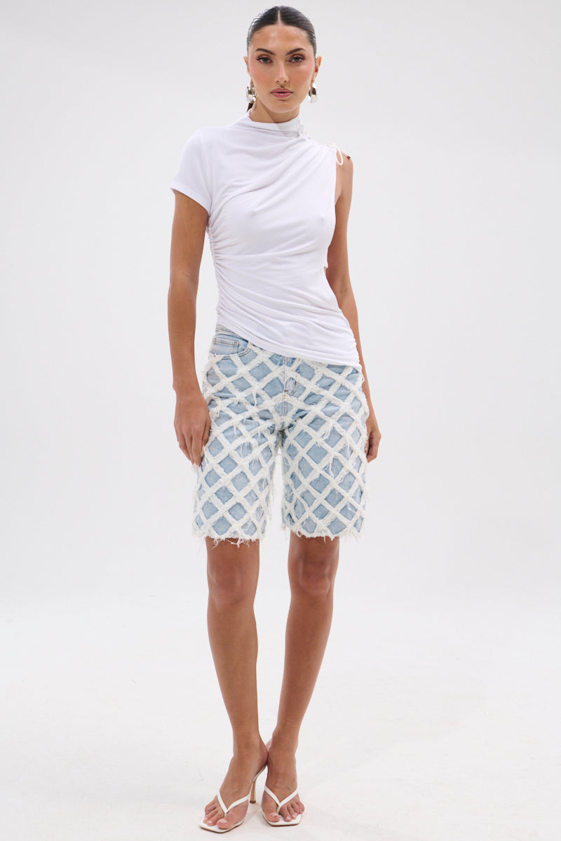 A woman stands against a white background wearing the SIMPLE AS THIS UNIQUE TEE, light blue knee-length denim shorts with a white lattice pattern, and white heeled sandals.