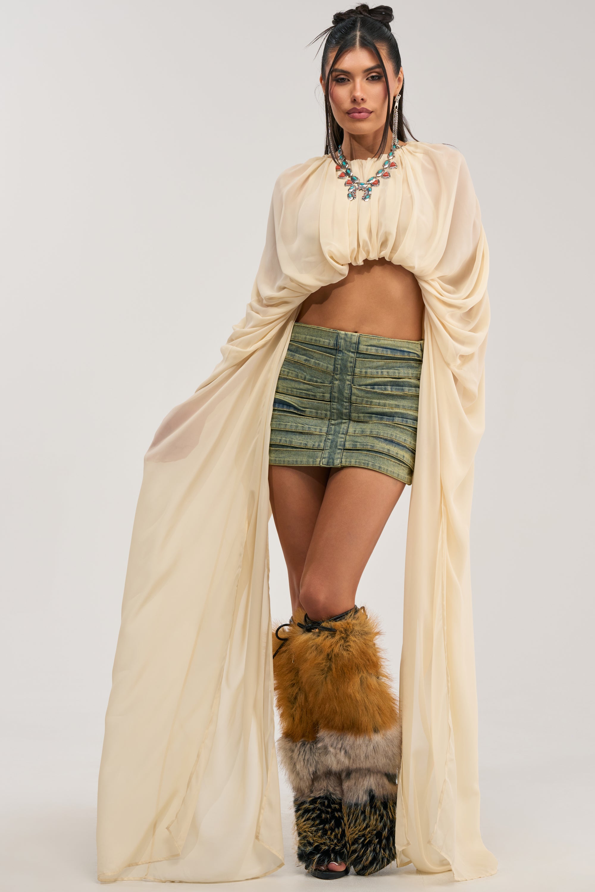 A woman poses in the FLY WITH ME CHIFFON BLOUSE IN IVORY, styled with a flowing beige cape, green striped mini skirt, statement necklace, and furry brown leg warmers against a plain light background.