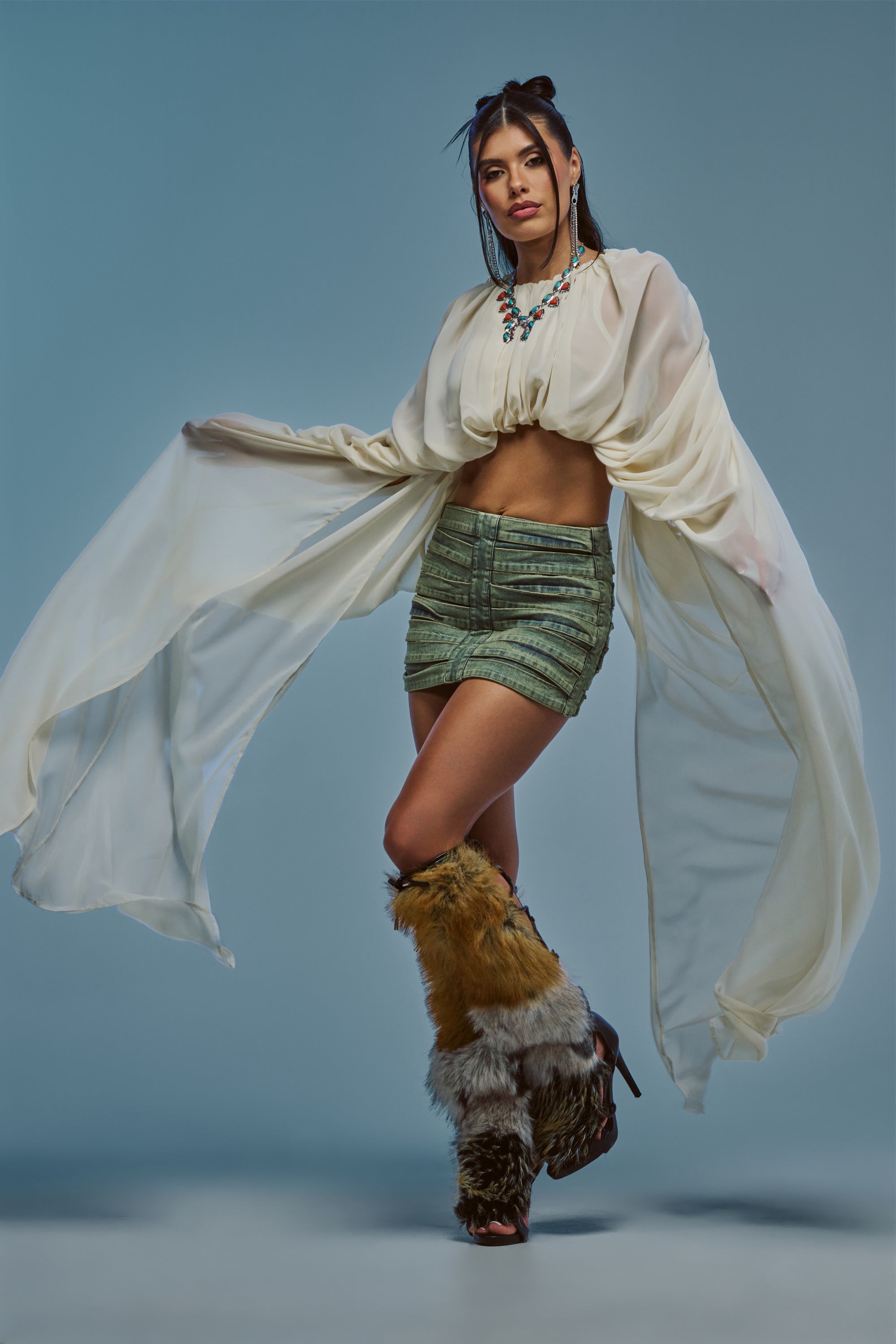 A woman poses in the FLY WITH ME CHIFFON BLOUSE IN IVORY with exaggerated sleeves, paired with green textured shorts, a statement necklace, and fur-trimmed boots, set against a blue background.