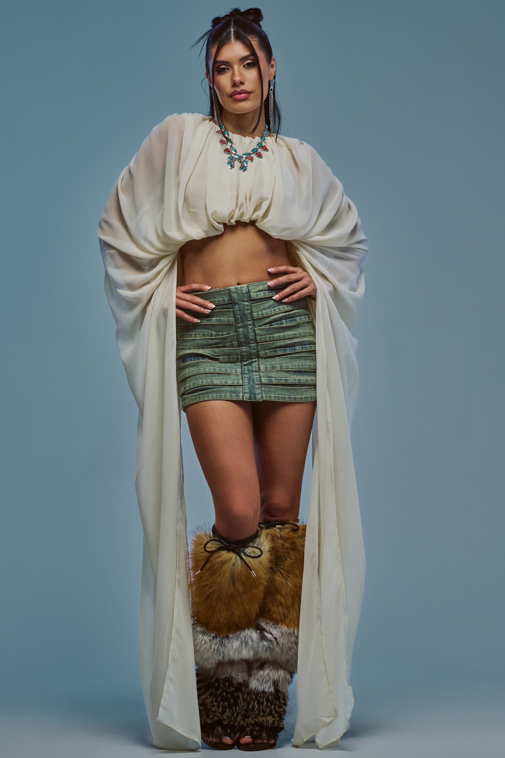 A woman poses in the FLY WITH ME CHIFFON BLOUSE IN IVORY, paired with a green mini skirt, brown furry boots, and a statement necklace, standing against a plain blue background.