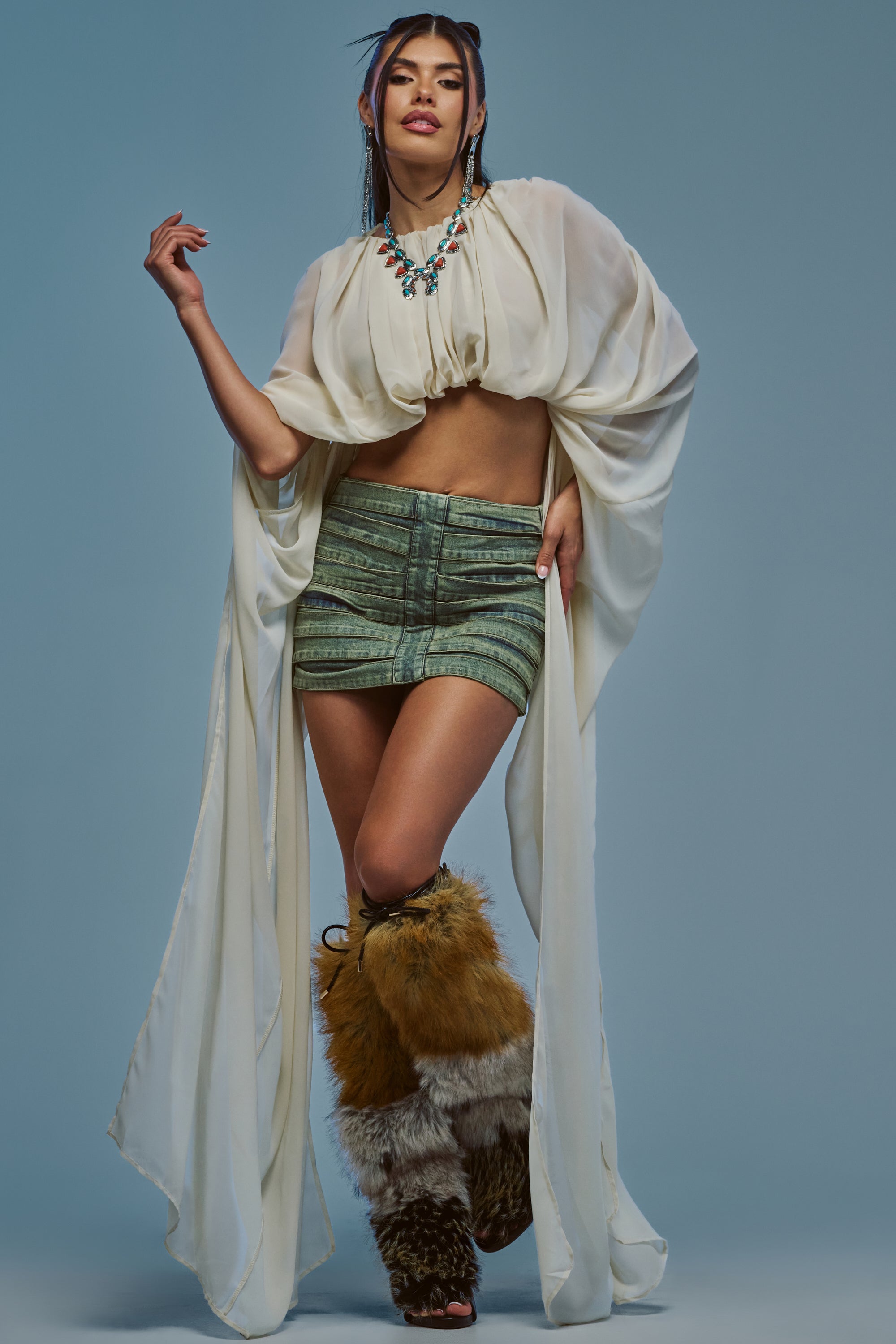 A woman poses in the FLY WITH ME CHIFFON BLOUSE IN IVORY, featuring exaggerated sleeves, paired with a green mini skirt, fur-covered boots, and a statement necklace, standing against a blue background.