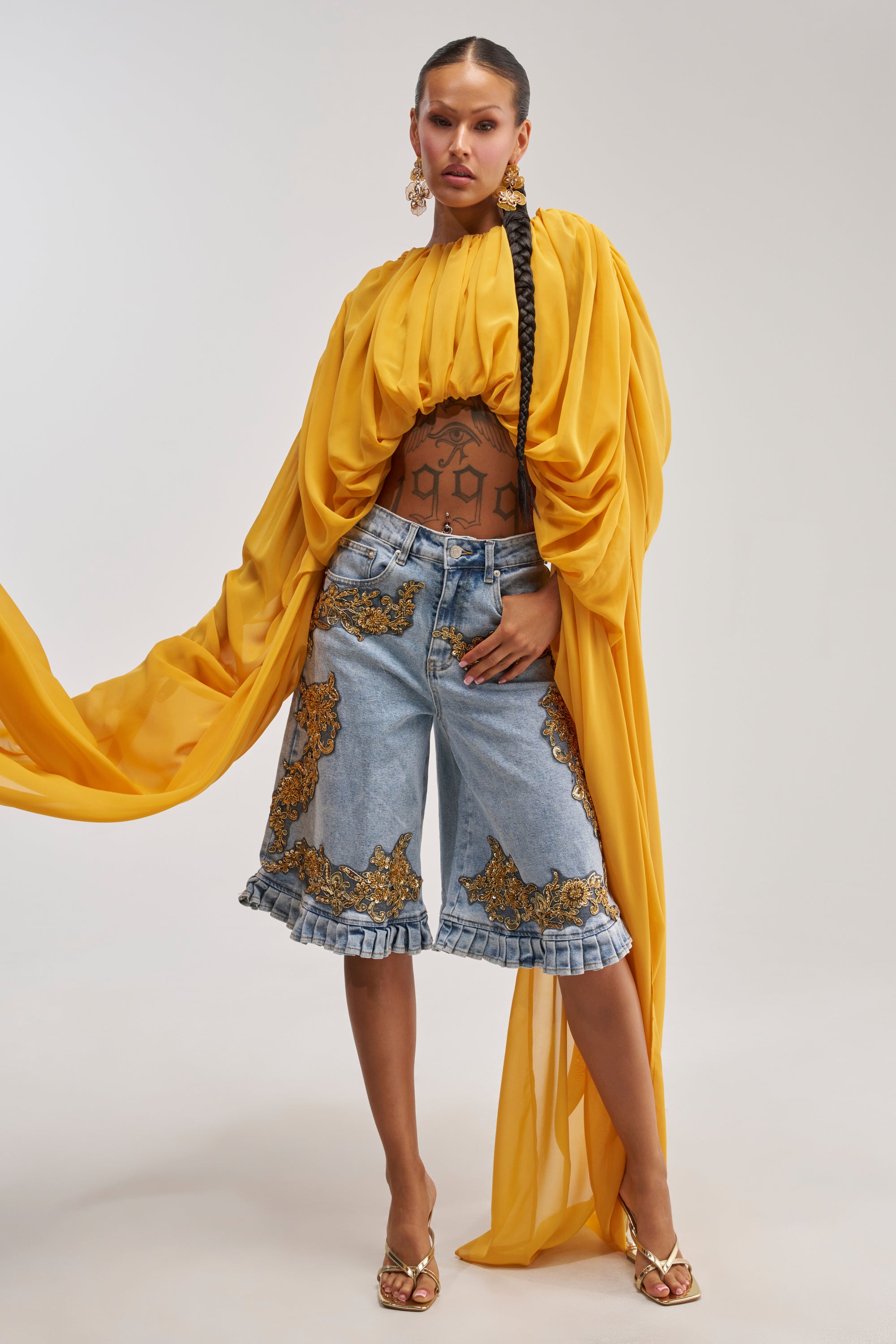 A woman poses in the FLY WITH ME CHIFFON BLOUSE—its flowing mustard yellow chiffon and draped sleeves styled with denim shorts featuring gold embroidery and ruffled hems, gold heels, hoop earrings, and a long braided ponytail for an effortlessly chic vacation look.