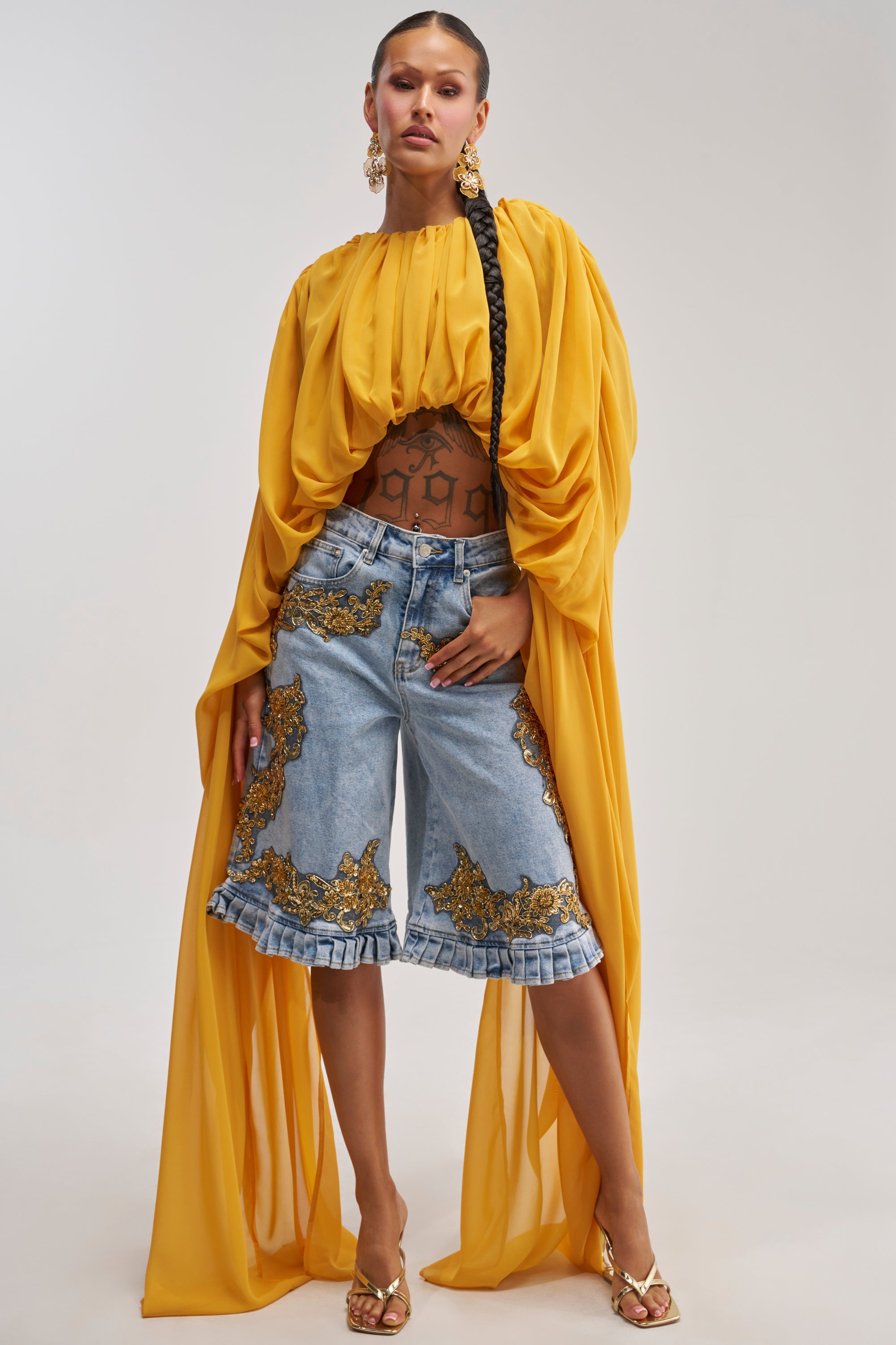 A woman showcases the FLY WITH ME CHIFFON BLOUSE in a vibrant mustard yellow, its flowing chiffon fabric and dramatic sleeves paired over denim shorts with gold accents. She completes her vacation-ready look with gold heels, statement earrings, a long braid, and visible tattoos for added flair.