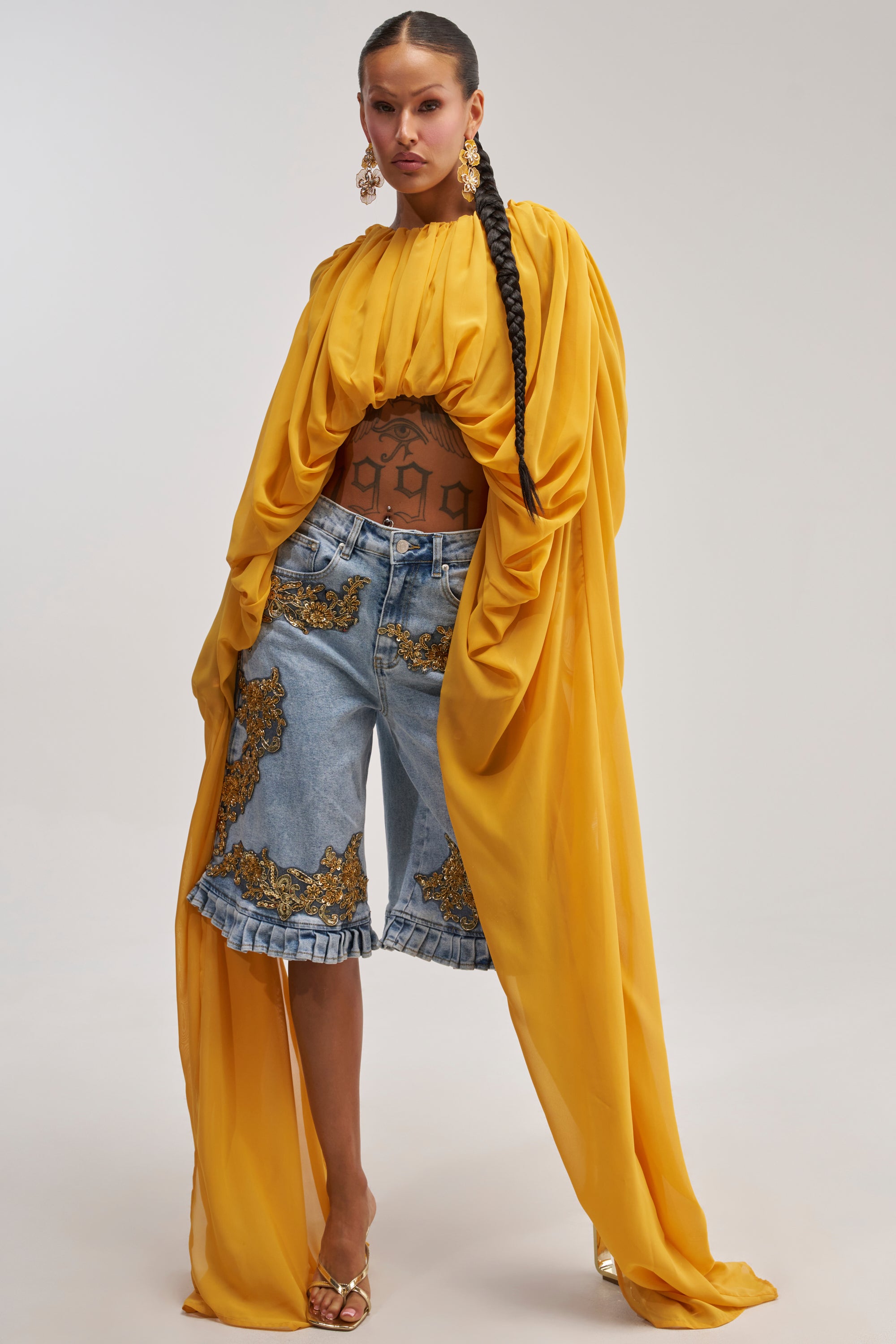 Dressed in the FLY WITH ME CHIFFON BLOUSE—a flowing mustard yellow top with long sleeves—she pairs it effortlessly with denim shorts featuring gold embellishments, gold heels, and statement earrings. Perfect for vacation styling, her look is finished with a long braid and visible tattoos on her midriff.