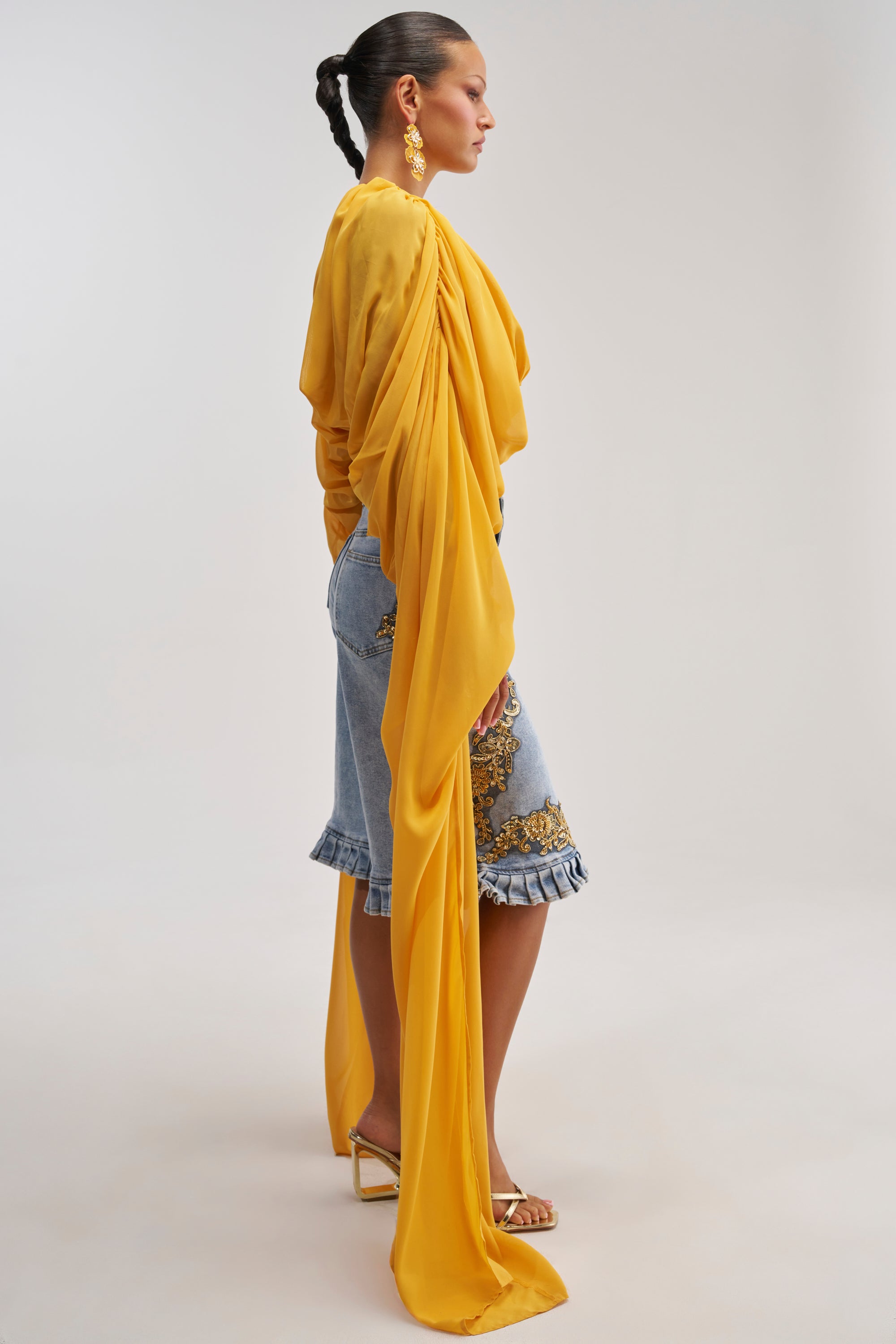 Woman in profile wearing the FLY WITH ME CHIFFON BLOUSE in mustard yellow with long flowing sleeves, paired with embroidered denim shorts featuring ruffles, gold earrings, and strappy heels—an effortless vacation-ready look—standing against a plain background.