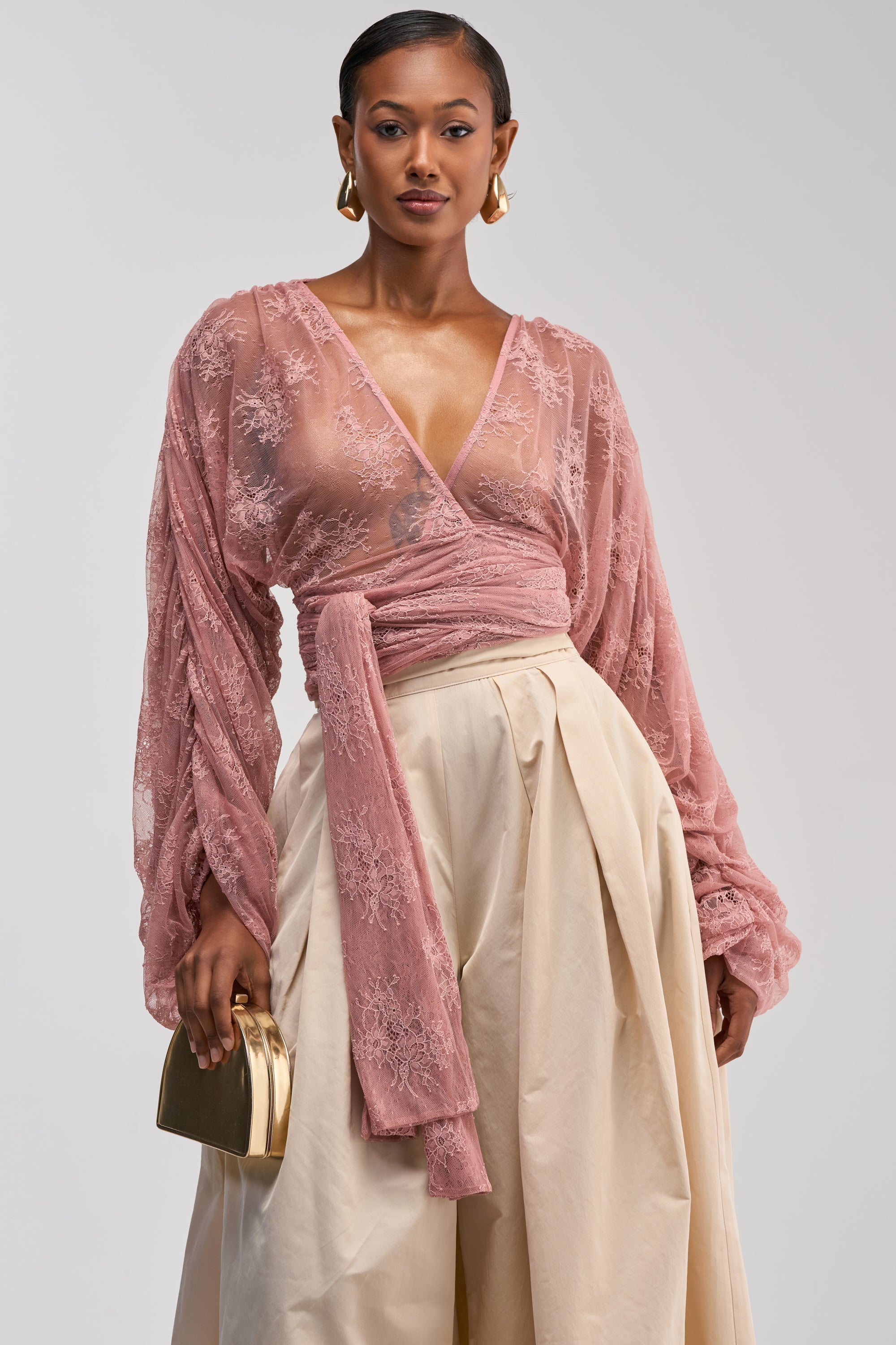 Wearing the LOCKED IN LOVE LACE BLOUSE—a sheer mauve lace wrap top with long sleeves—paired with cream high-waisted wide-leg pants, gold hoop earrings, and a small gold purse against a plain background, she creates an effortlessly elegant look that’s perfect for a romantic date night.