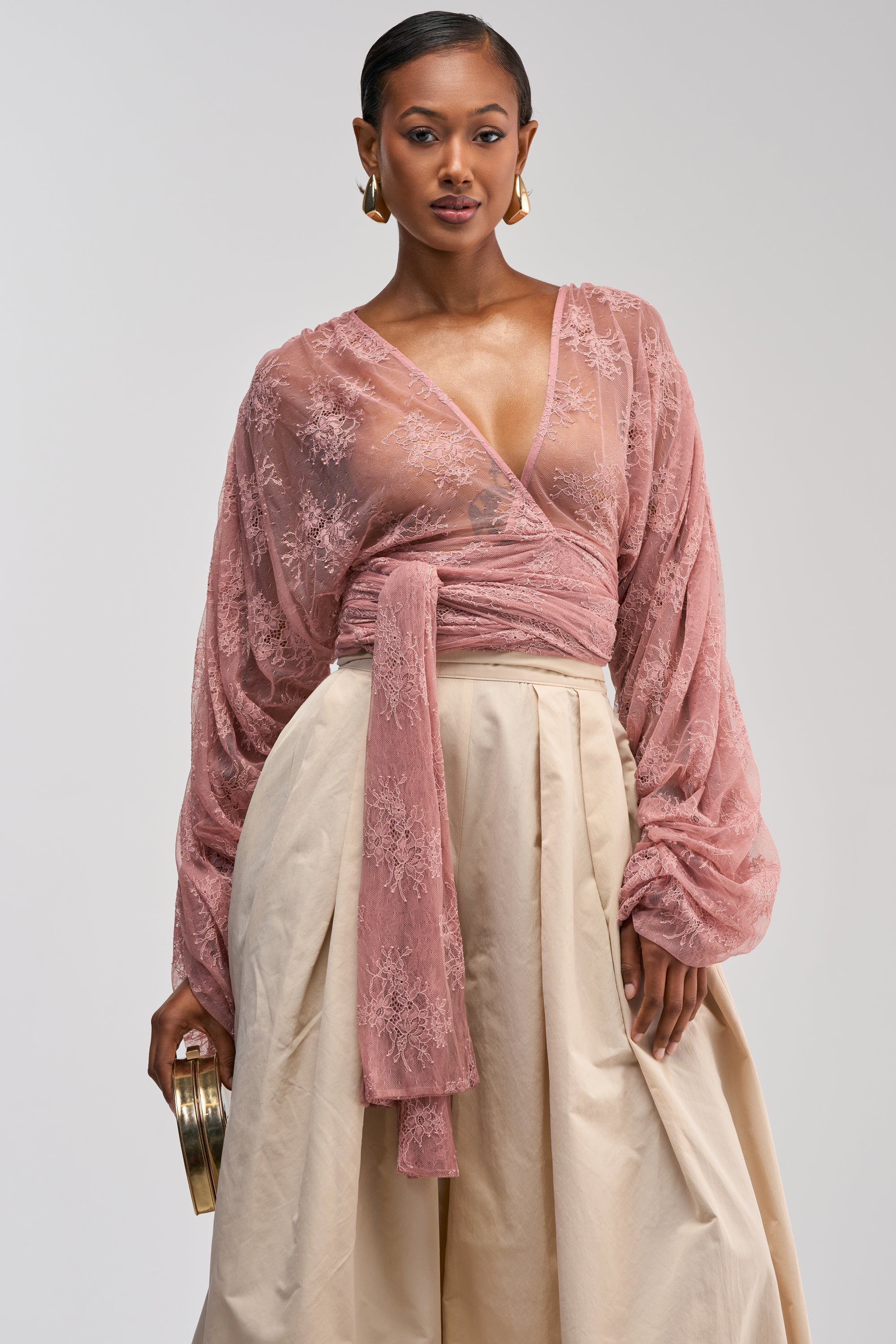 Wearing the LOCKED IN LOVE LACE BLOUSE—a sheer, mauve lace wrap top with long, voluminous sleeves that’s perfect for a romantic date night—a woman pairs it with a beige high-waisted skirt and holds a small gold clutch while posing against a plain background.