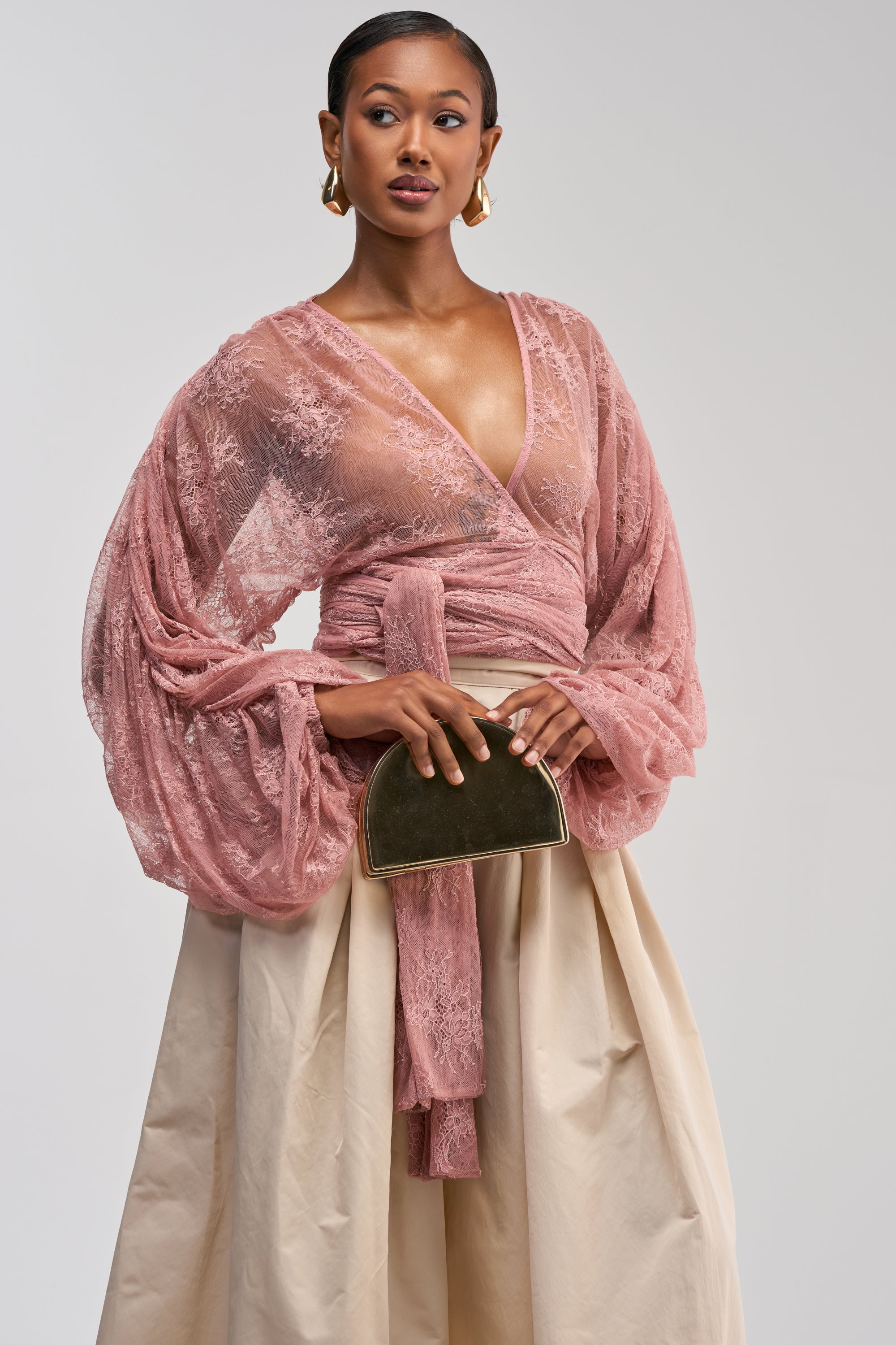 A woman wears the LOCKED IN LOVE LACE BLOUSE—a sheer pink wrap top with billowy sleeves—styled elegantly with a cream skirt and a small, dark green clutch. Gold hoop earrings complete her look as she stands against a plain light background.