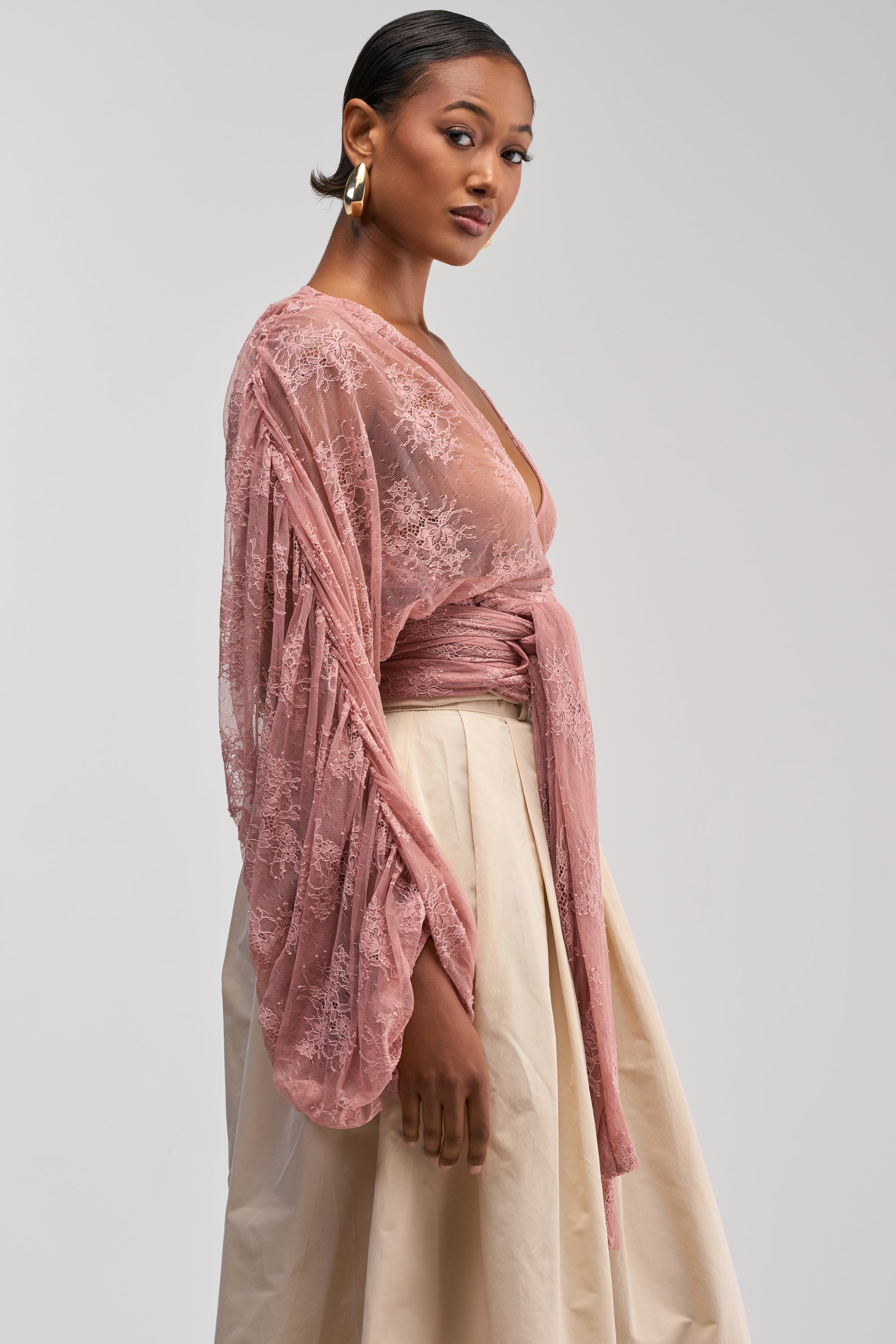 Wearing the LOCKED IN LOVE LACE BLOUSE—a sheer pink lace wrap top with wide sleeves—and a cream-colored skirt, a woman stands against a plain light background, looking confidently at the camera.