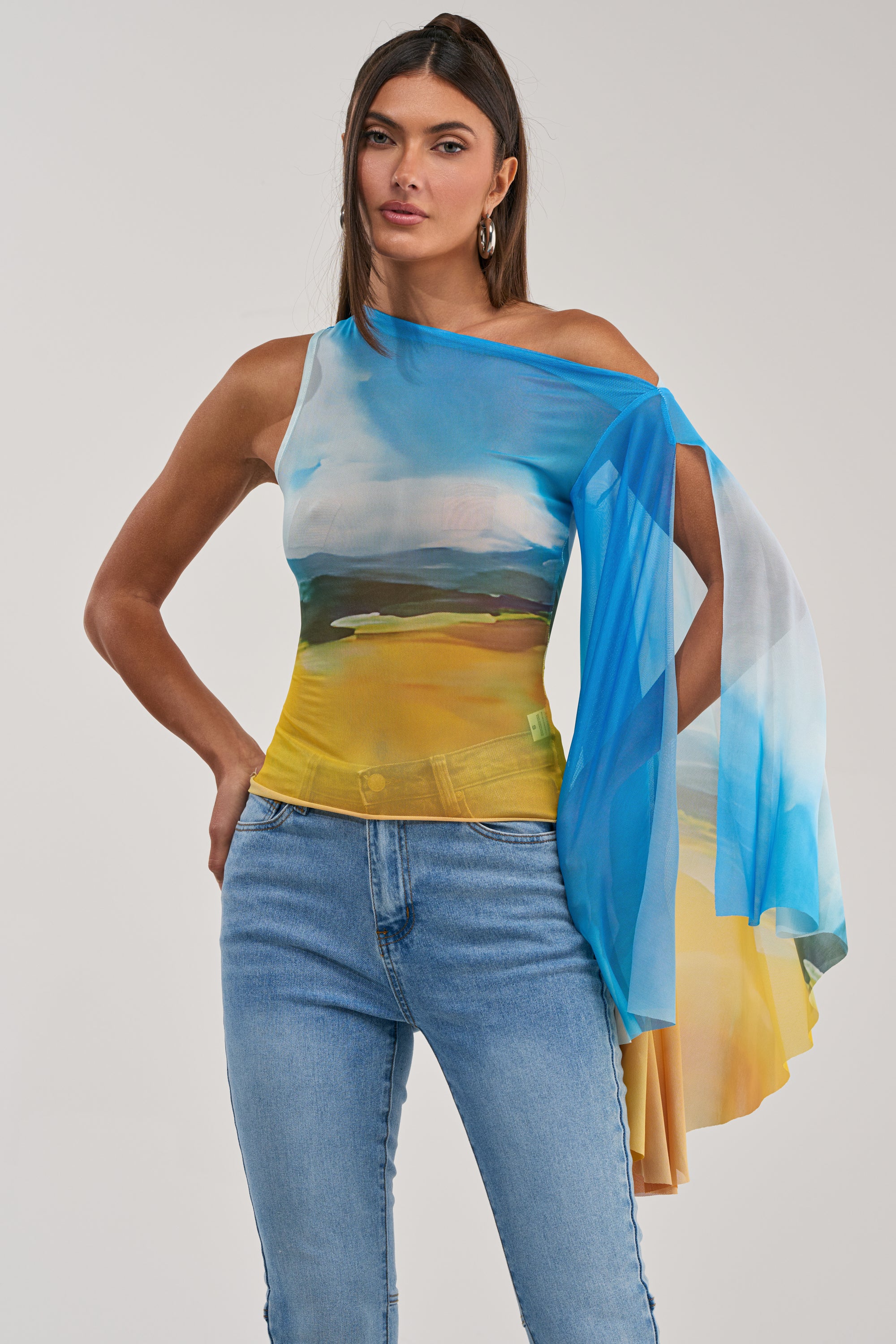 A woman models the PICTURESQUE LONG SLEEVE MESH TOP in blue, white, and yellow sky print with light blue jeans, standing with one hand on her hip against a plain background.