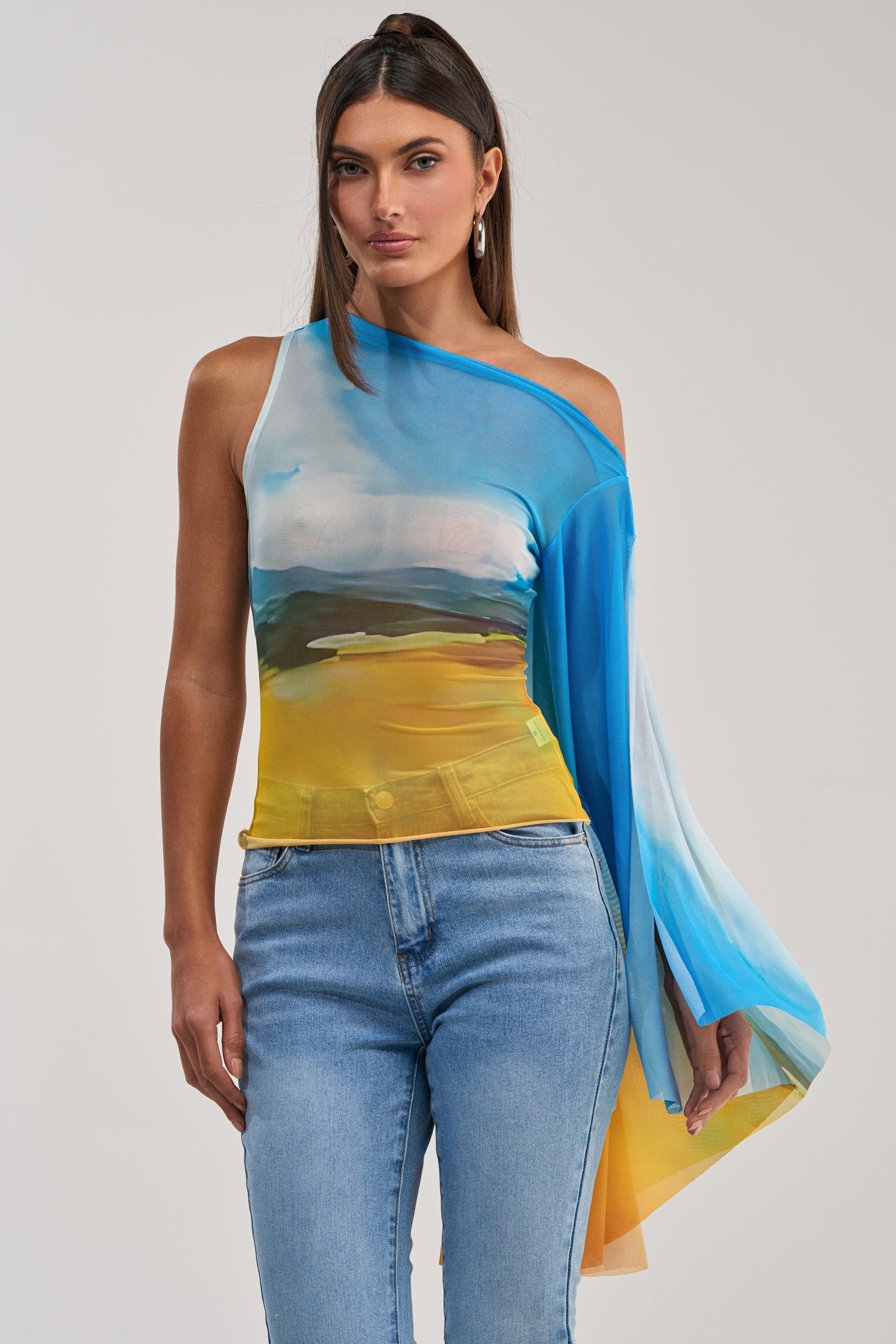 A woman wears the PICTURESQUE LONG SLEEVE MESH TOP with a blue, yellow, and green landscape print and flowing sleeve, paired with light blue jeans, standing against a plain light background.