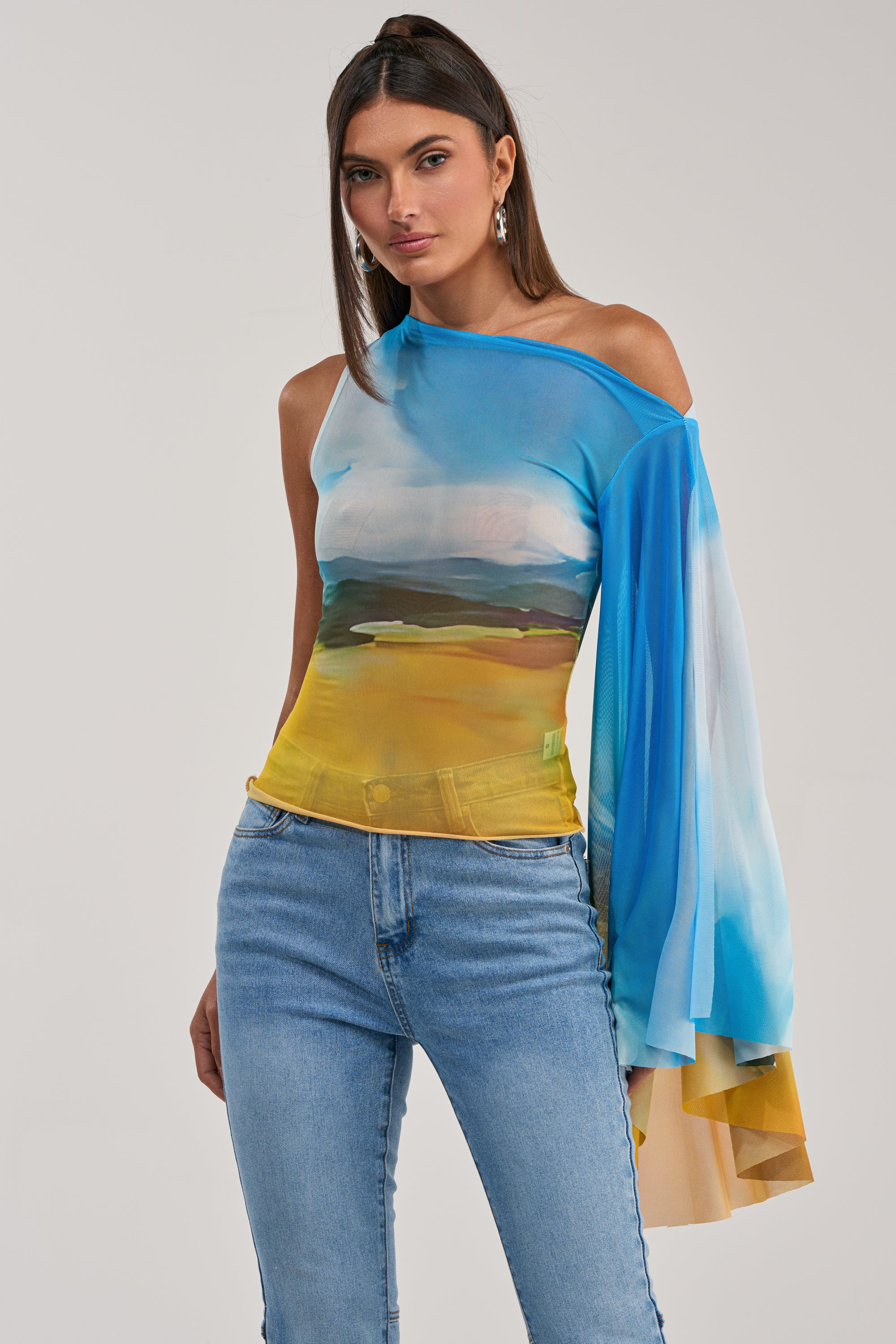 A woman poses in the PICTURESQUE LONG SLEEVE MESH TOP with a landscape print, featuring one sheer sleeve and an off-the-shoulder design, paired with blue jeans. The perfect standout summer party top.