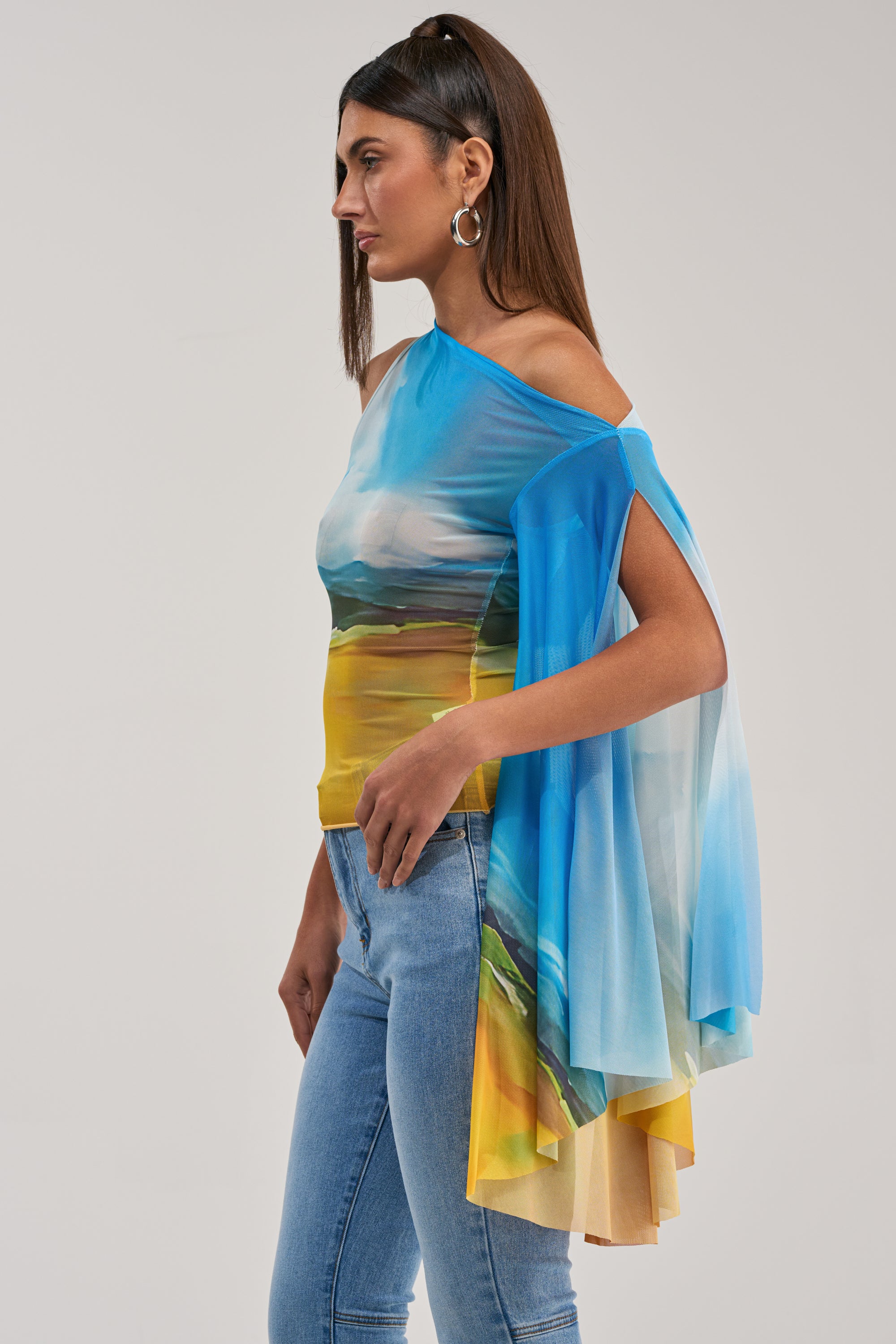 A woman in profile wears the PICTURESQUE LONG SLEEVE MESH TOP with blue, yellow, and green tones, light blue jeans, gold hoop earrings, and long straight hair. The top features a sheer mesh one-sleeve cape for a vibrant party look.