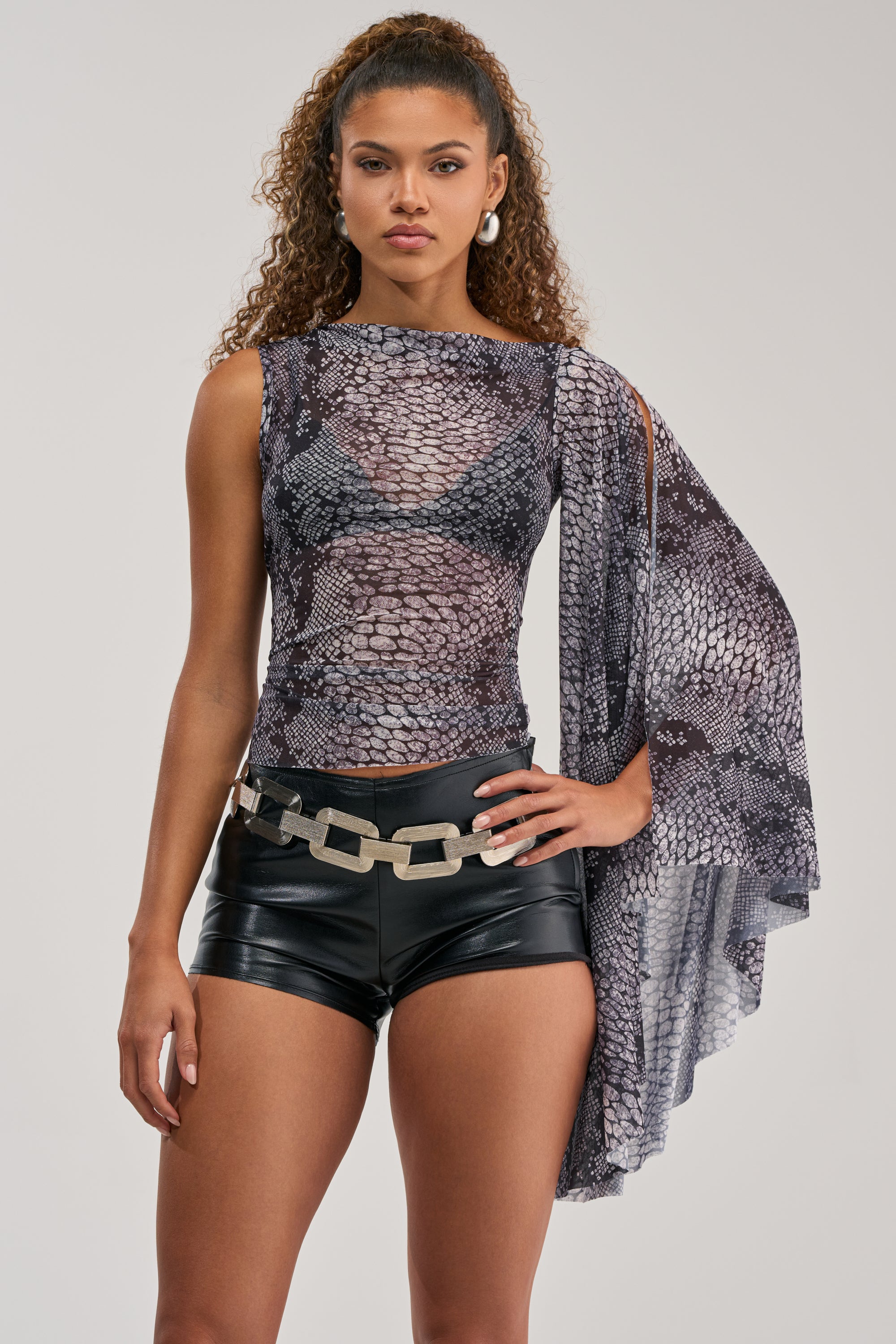 A woman stands facing forward against a plain background, wearing the SNAKEBITE ONE-SLEEVE TOP—a mesh, snakeskin-patterned piece—paired with black leather shorts, a chunky metallic belt, and hoop earrings.