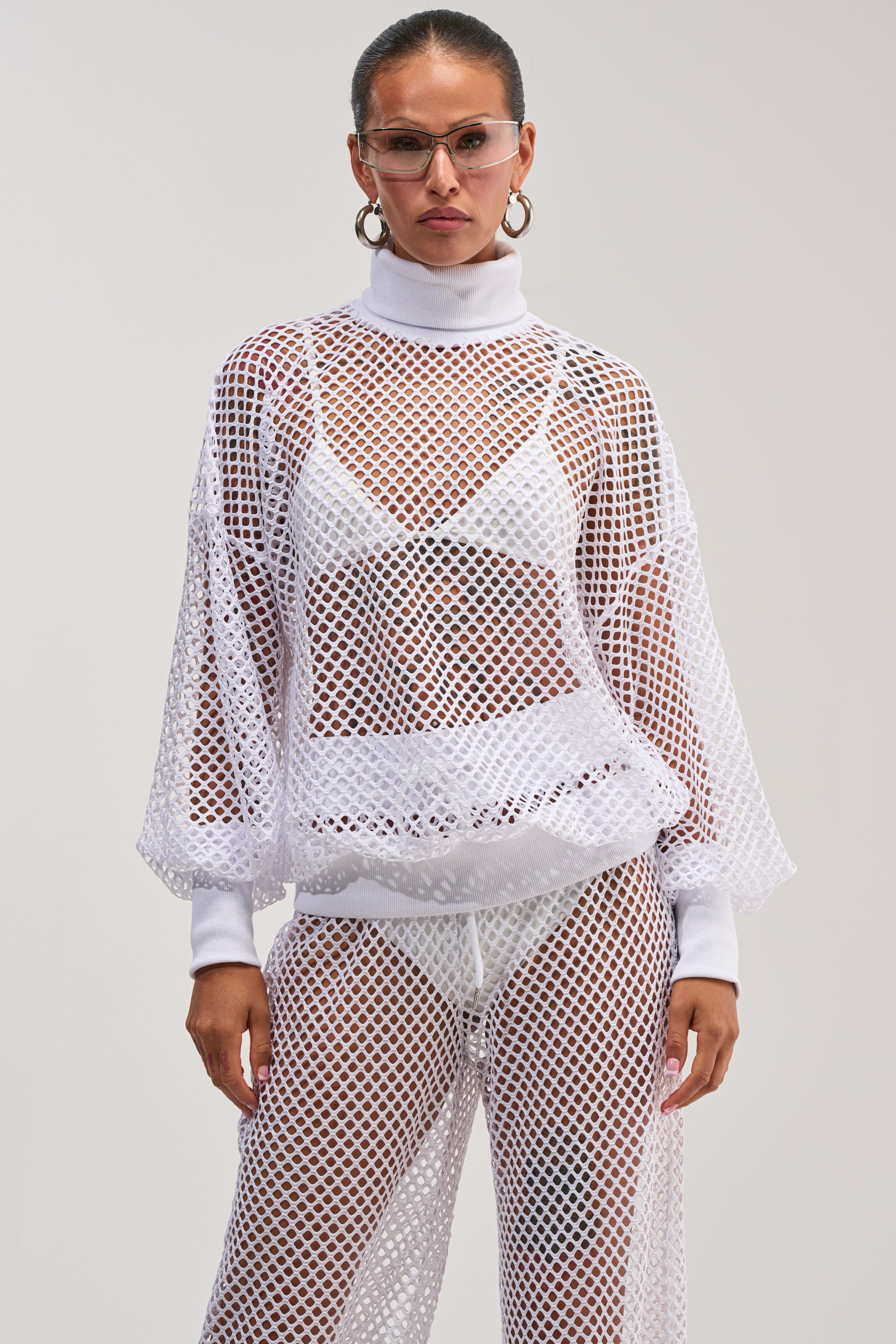 Wearing the FEEL THE AIR NETTED LONG SLEEVE layered over a white bra and pants, accessorized with large hoop earrings and glasses, this look against a plain background serves up fresh inspiration for standout beach day style.
