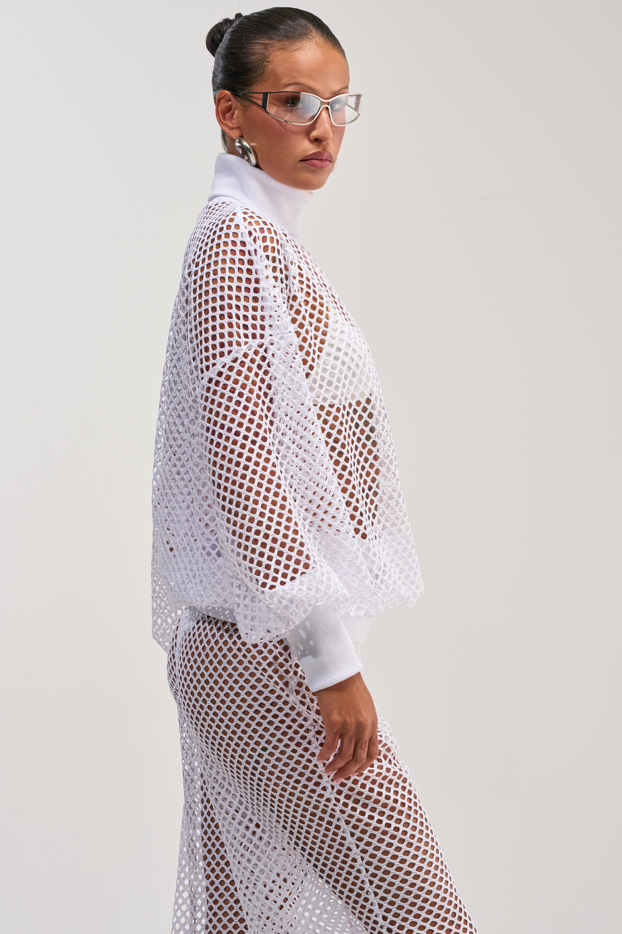A woman poses against a plain light background wearing the FEEL THE AIR NETTED LONG SLEEVE, styled with glasses and her hair in a bun.