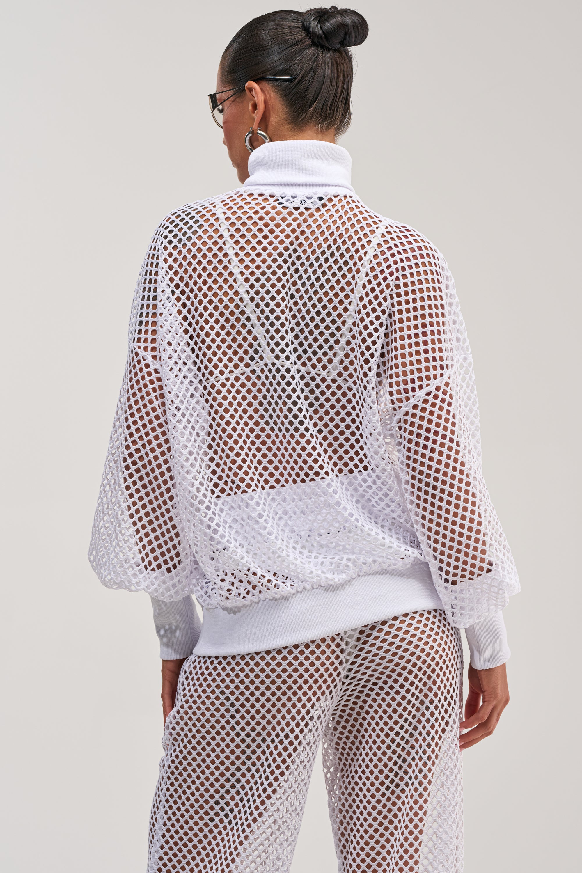A woman with her hair in a bun stands with her back to the camera against a light background, wearing the FEEL THE AIR NETTED LONG SLEEVE and matching pants set over a white bra and briefs. This see-through ensemble captures an effortless beach day vibe with its airy, stylish design.