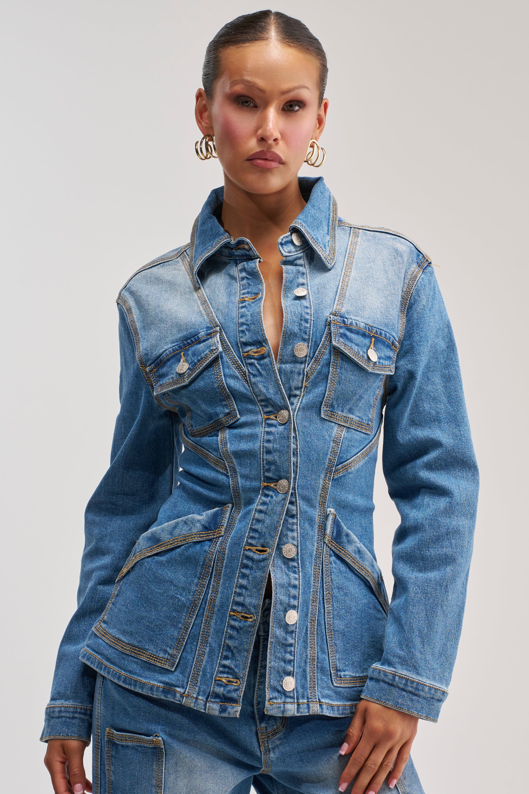 Wearing the DENIM FOR DAYS BUTTON DOWN—a fitted blue denim jacket with silver buttons—paired with matching denim jeans, a woman embodies classic denim on denim streetwear style against a simple light background.