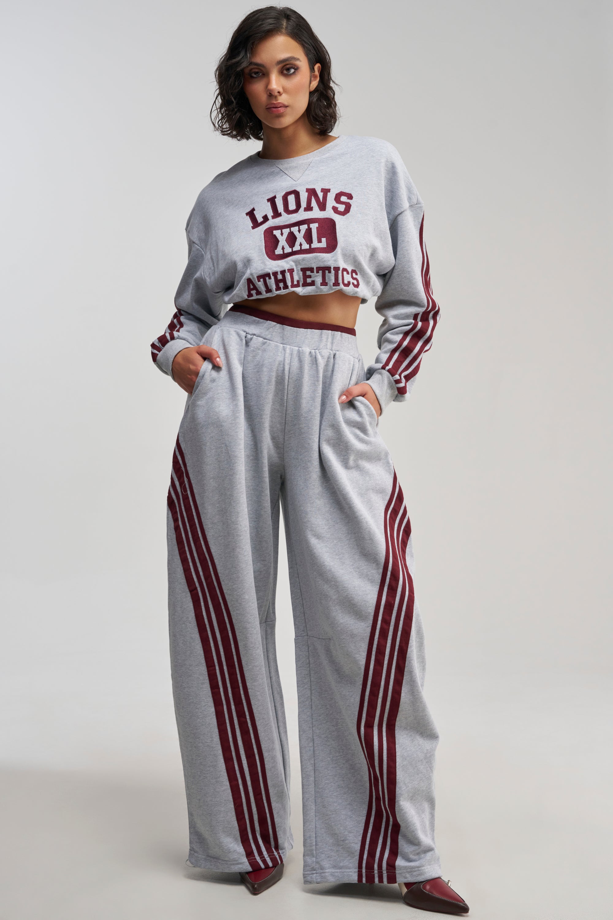A woman stands against a neutral background wearing the Victory Lane Striped Jogger in heather grey fleece, paired with a matching cropped sweatshirt featuring maroon stripes and “LIONS XXL ATHLETICS,” hands in pockets, completing this athleisure look with wide-leg sweatpants.