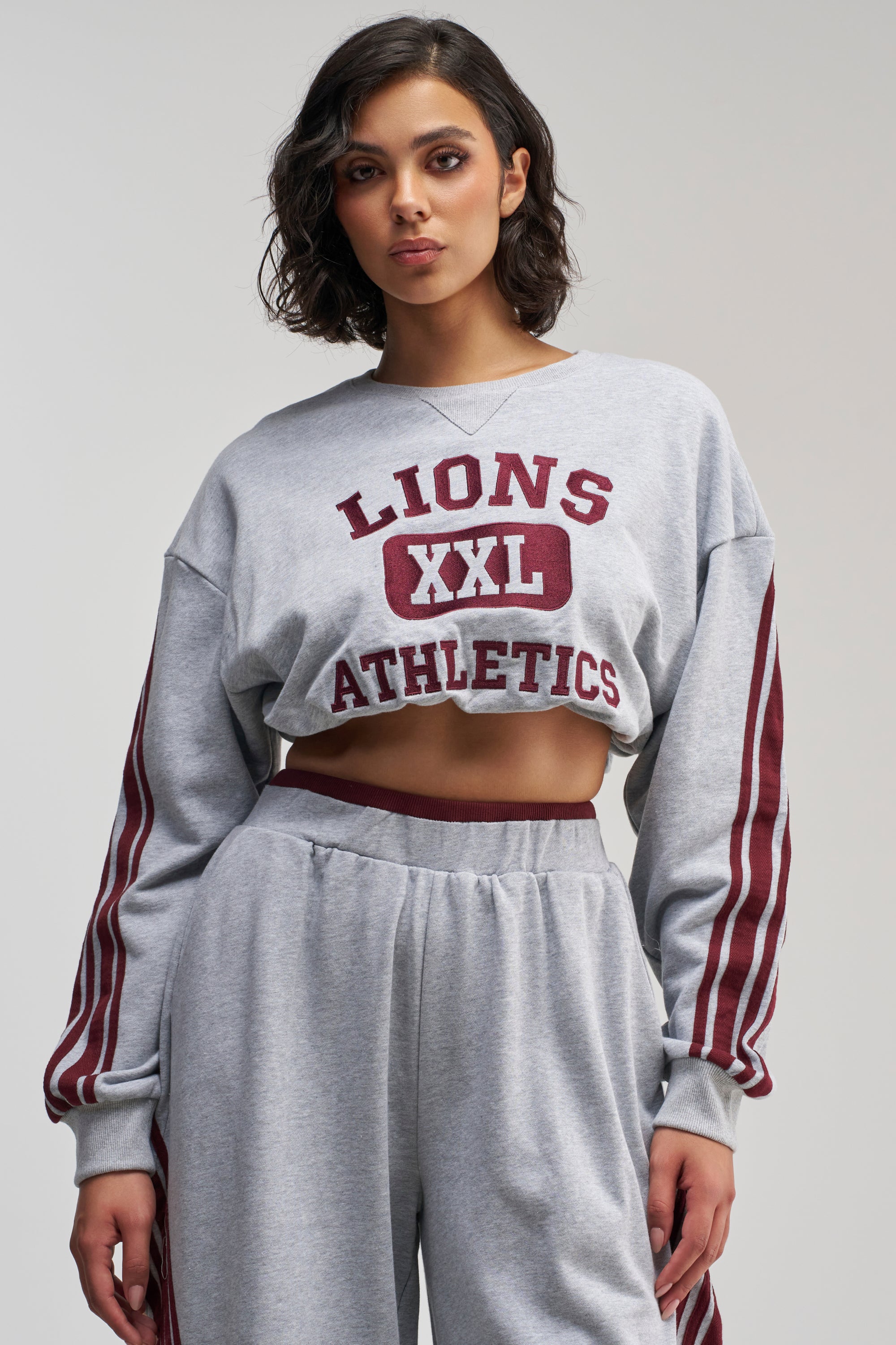 A woman stands against a plain background, wearing the VICTORY LANE STRIPED SWEATSHIRT—a grey varsity-style top featuring “LIONS XXL ATHLETICS” in maroon text—paired with matching sweatpants with maroon stripes, showcasing a sporty, oversized street-style silhouette.