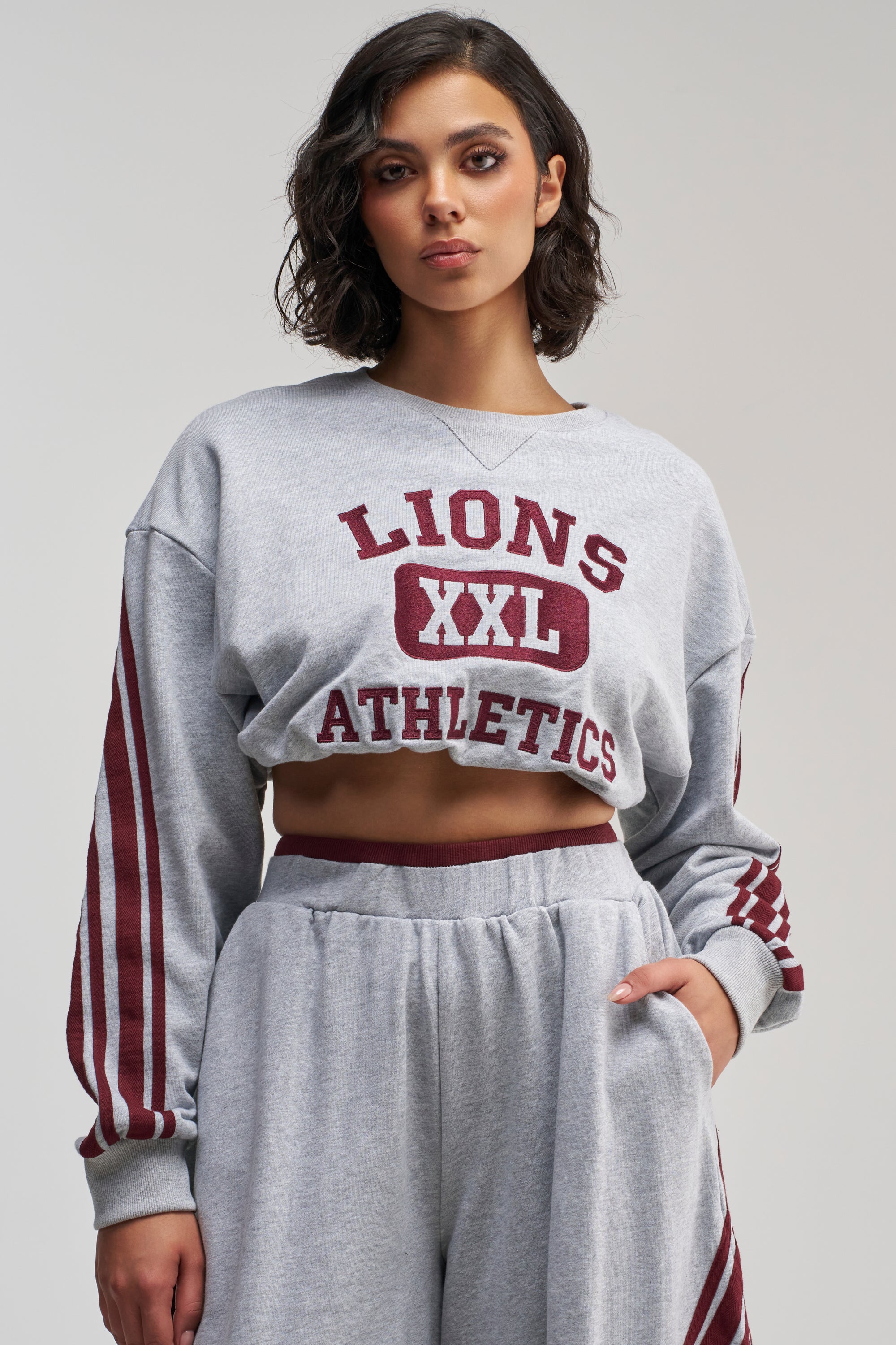 Wearing the VICTORY LANE STRIPED SWEATSHIRT in grey, featuring “Lions XXL Athletics” in maroon text, paired with matching sweatpants accented by maroon stripes, a person stands with one hand in their pocket, effortlessly channeling a sporty street-style vibe.