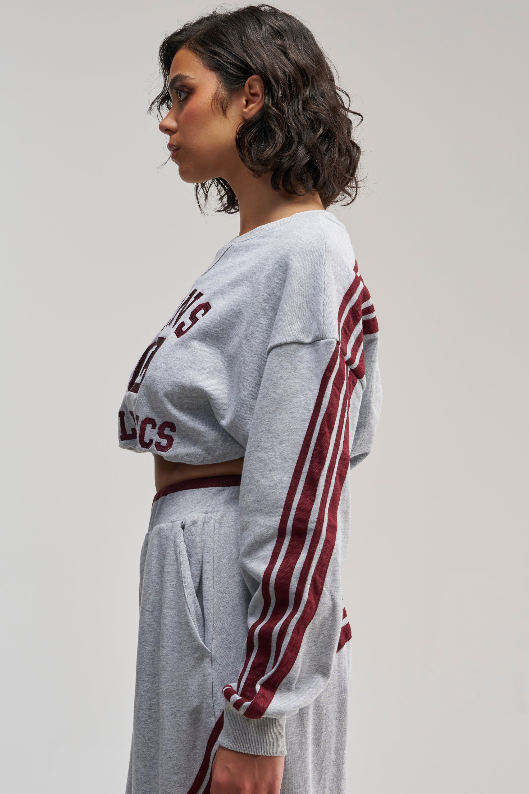 A woman in profile showcases the VICTORY LANE STRIPED SWEATSHIRT in grey, styled in an oversized silhouette and paired with matching sweatpants featuring maroon stripes and lettering, evoking a sporty street-style vibe against a plain light background.