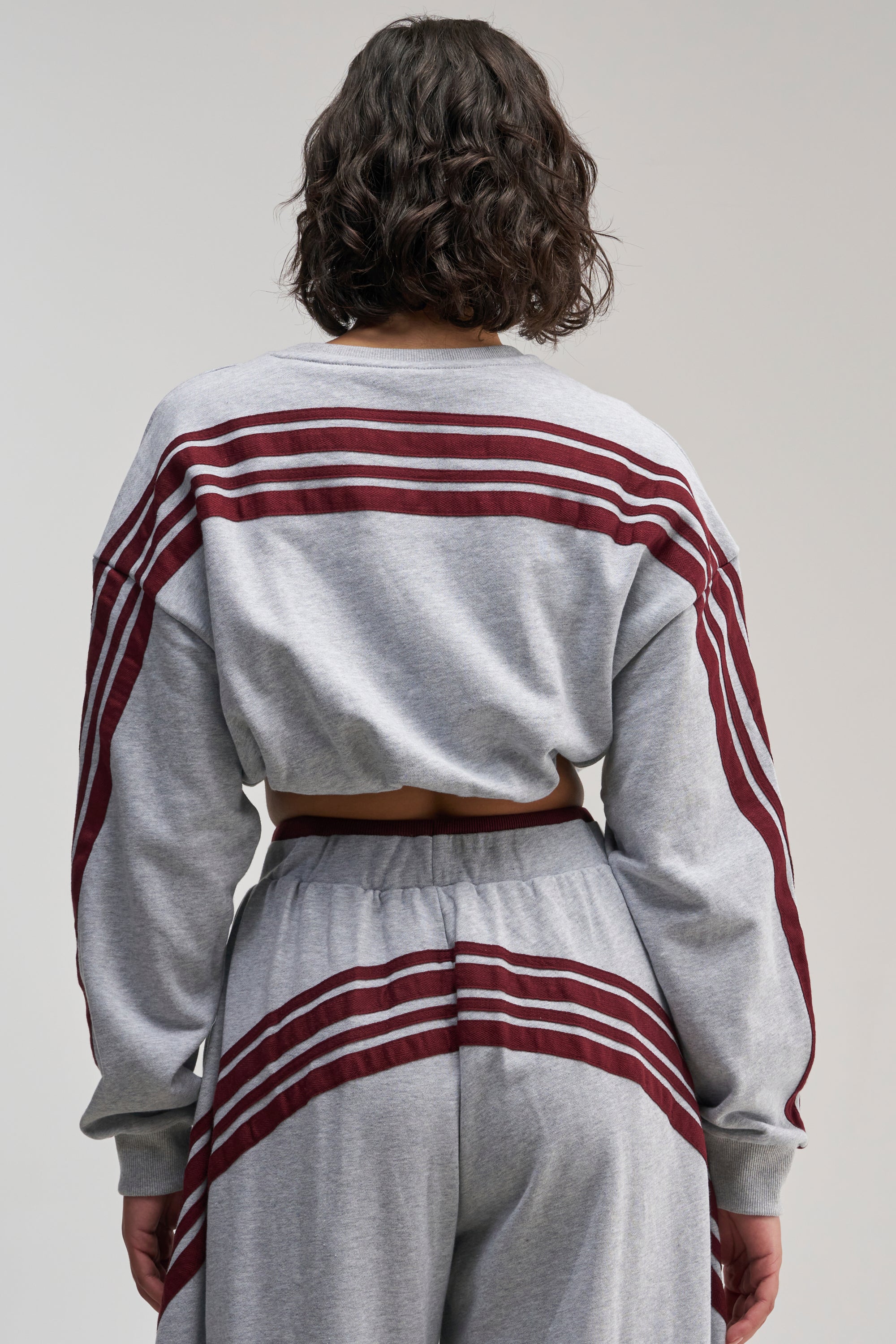Wearing the VICTORY LANE STRIPED SWEATSHIRT in an oversized fit, a person with wavy brown hair is seen from behind, showcasing maroon stripes along the sleeves, back, and matching pants against a simple light background for a sporty street-style vibe.