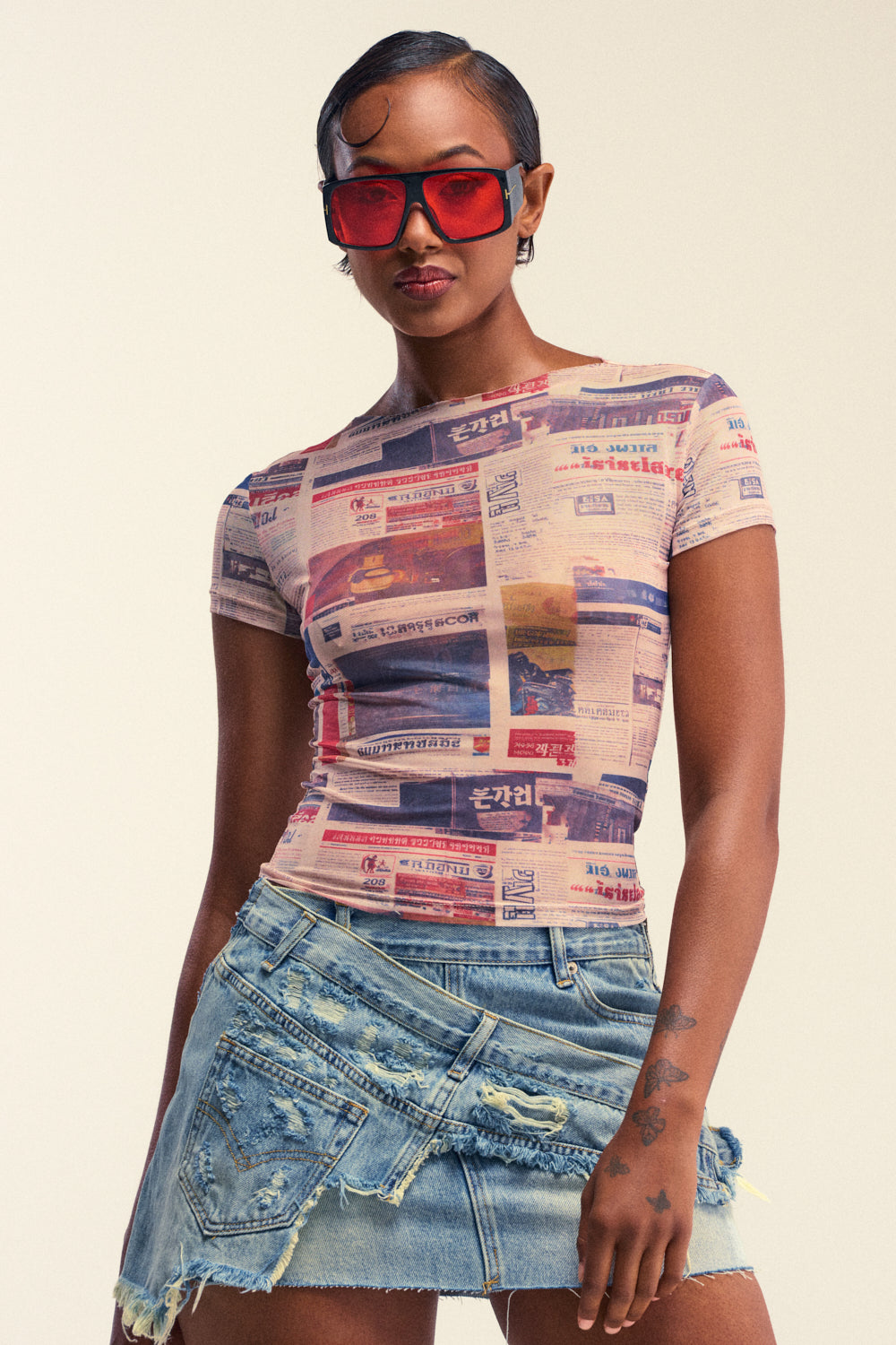A woman stands confidently against a neutral background, wearing red sunglasses, a distressed denim mini skirt, and the EXTRA EXTRA PRINTED MESH TEE—a fitted graphic newspaper print top that delivers an effortlessly bold statement streetwear look.