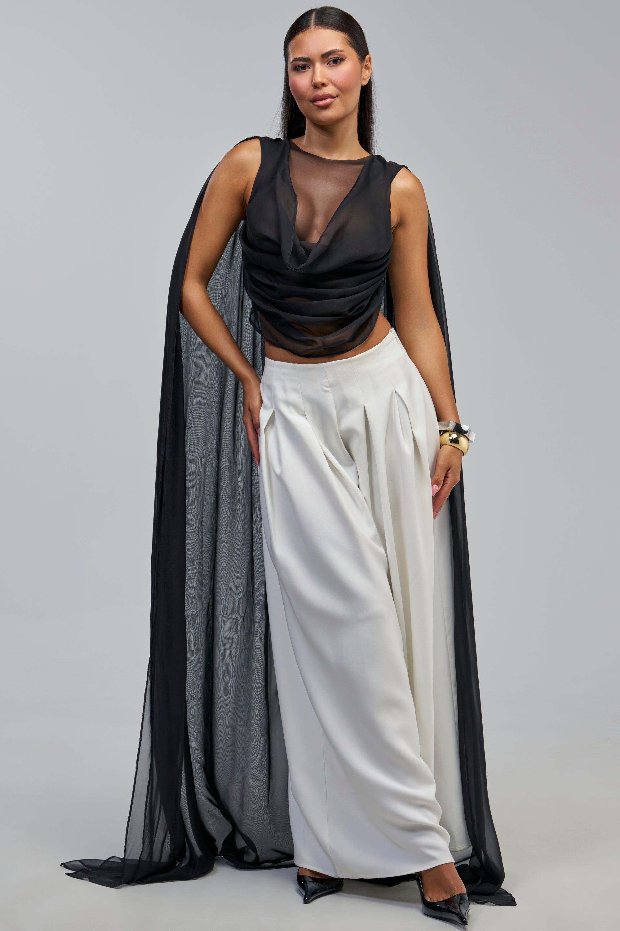 A woman dressed in the ANGELIC CHIFFON BLOUSE, paired with white wide-leg pants, black heels, and bracelets, stands against a plain gray background.