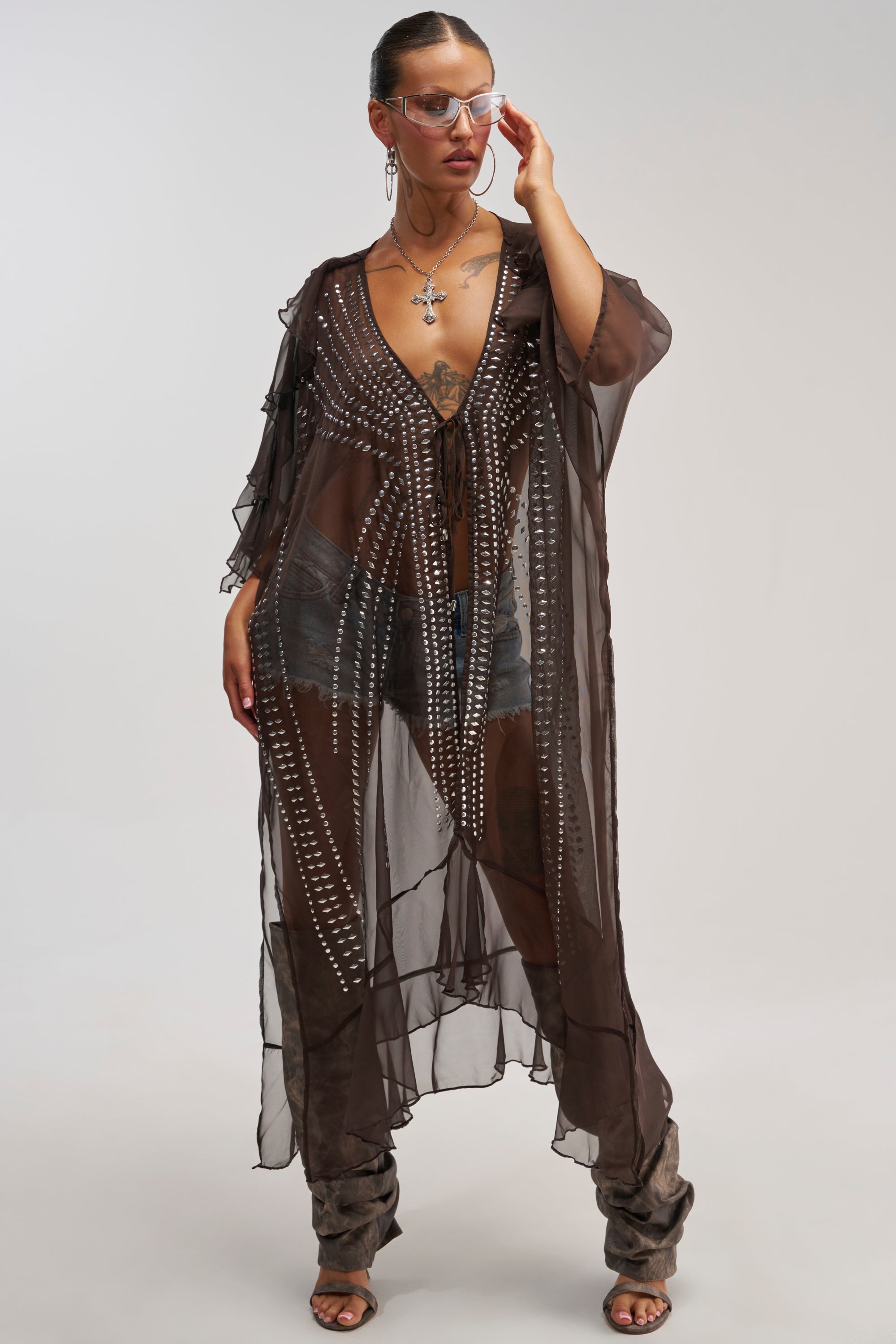 Dressed in the INDIO VIBES TIE FRONT CARDIGAN—a sheer, long brown chiffon duster adorned with silver embellishments—she layers it over a black bra and shorts. Tall slouchy boots and sunglasses complete her confident look against a plain background, creating a striking festival outfit that exudes effortless style.