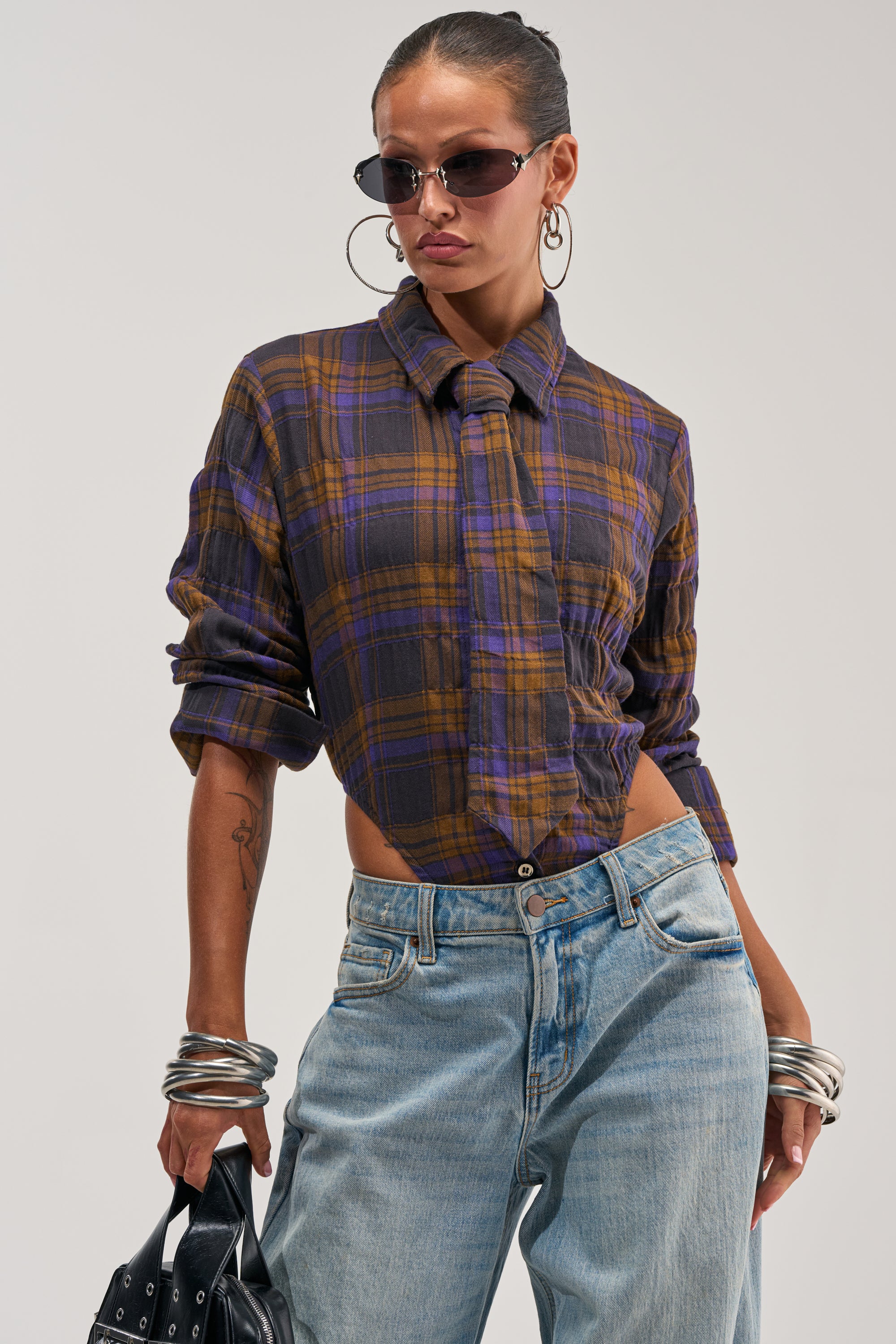 Wearing the NOW YOU SEE ME PLAID BODYSUIT tied at the waist with loose jeans, bold sunglasses, and large hoop earrings, she accessorizes with a black bag and chunky bracelets, striking a confident pose against a plain background for cozy-chic styling.