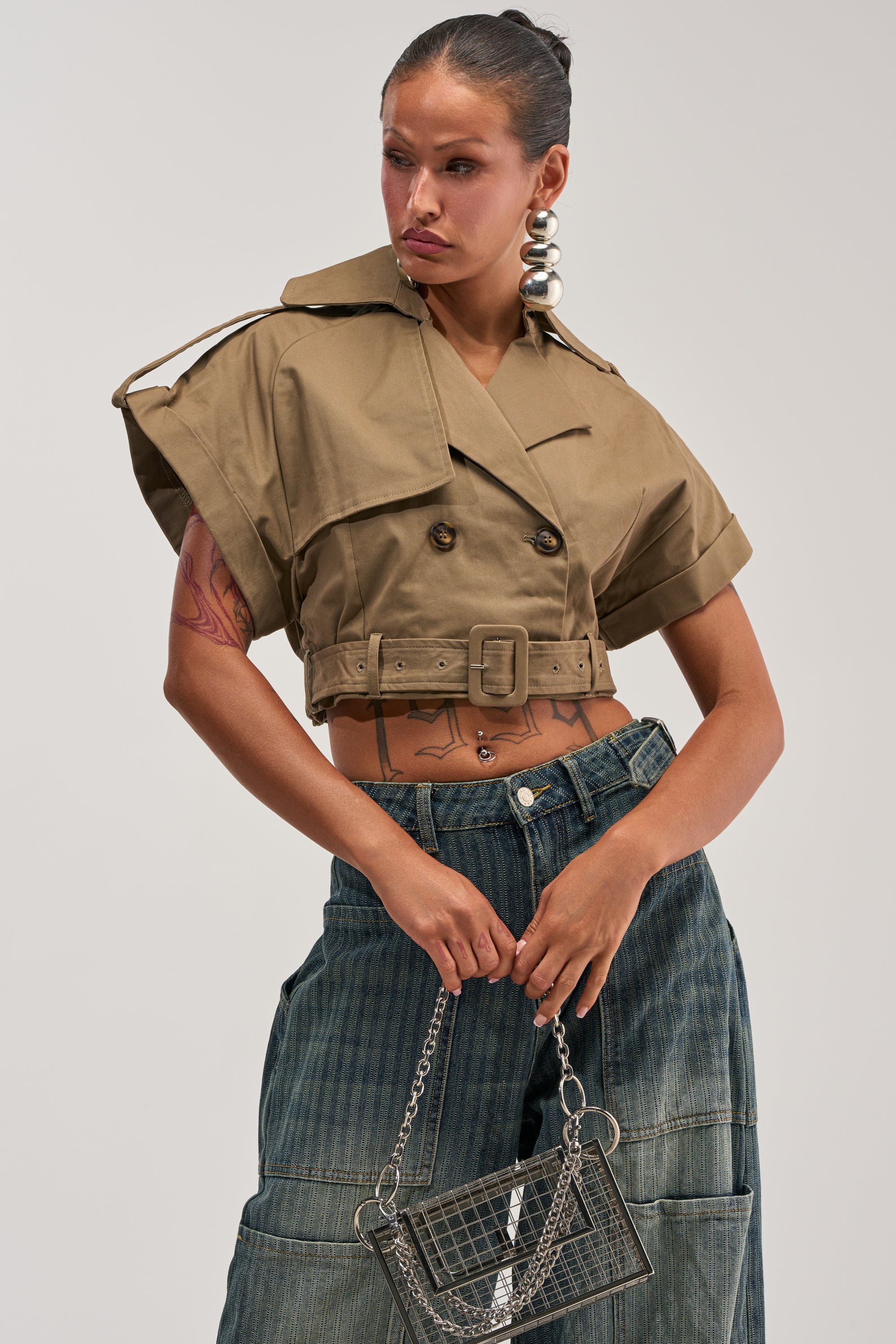 A woman wearing the OUT AND ABOUT CROPPED TRENCH TOP in khaki, paired with wide dark denim pants, large silver earrings, and holding a metallic purse with a chain strap. She has visible tattoos and a confident expression.