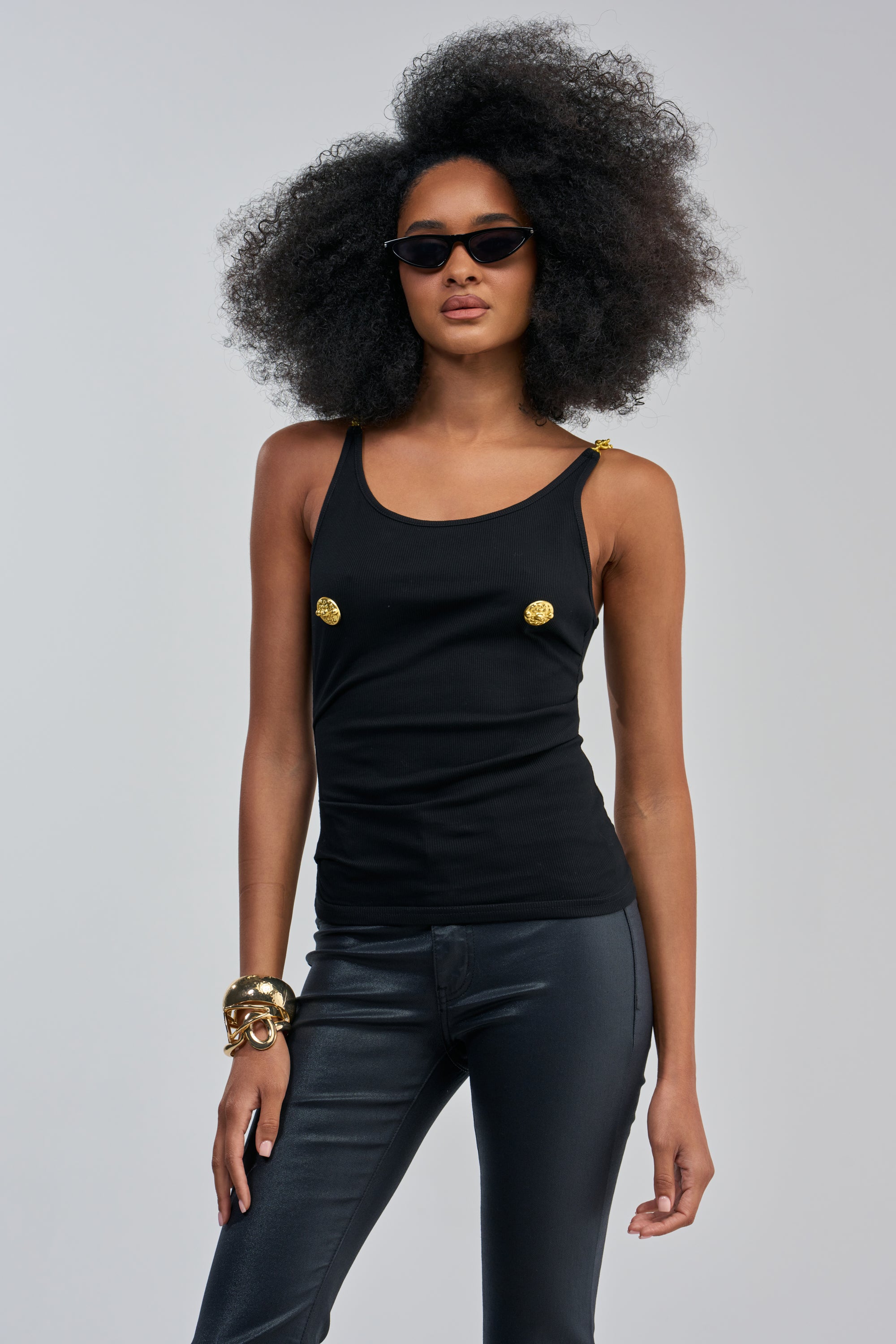 Woman with voluminous curly hair in black sunglasses, wearing the EYES ON ME KNIT TANK paired with black pants, accessorized with gold bracelets and a chic gold chain strap, standing against a plain background.