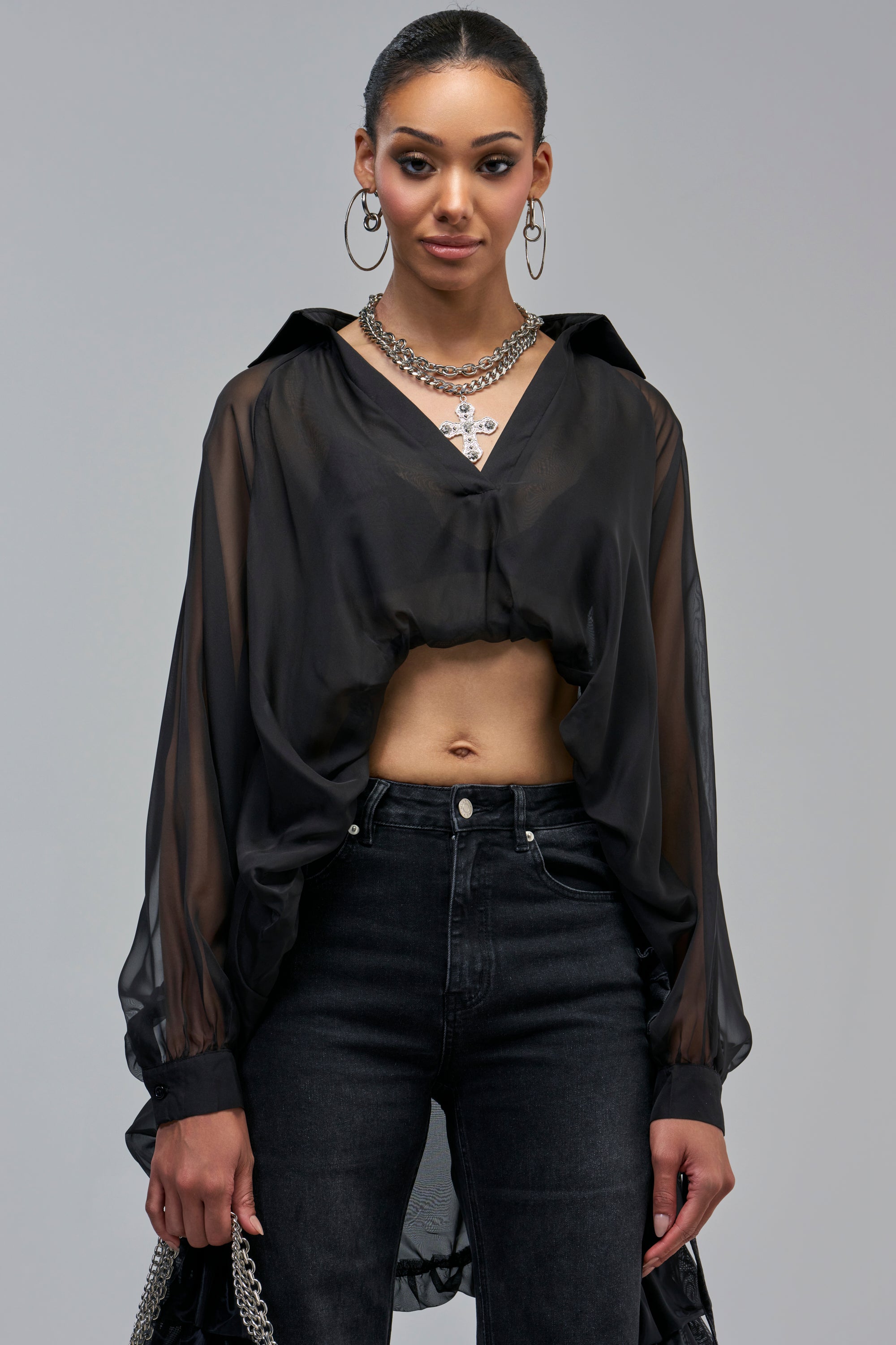 A person wears the SHEER PRESSURE CHIFFON BUBBLE HEM TOP IN BLACK with dark jeans and a layered silver chain necklace featuring a cross pendant.