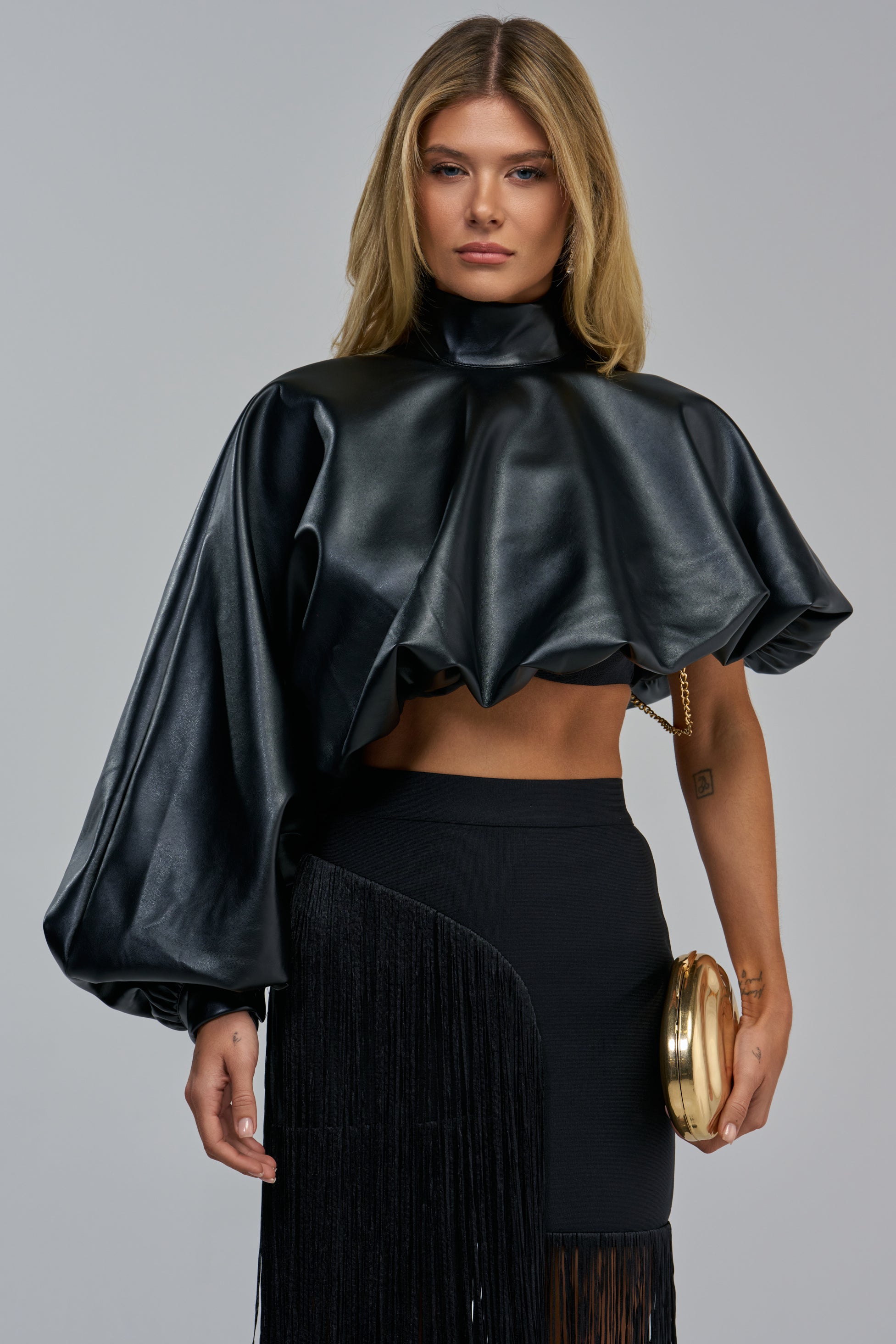 A woman wears an avant-garde YOU WOULDNT GET IT FAUX LEATHER BLOUSE with a high neck, voluminous sleeves, and a black fringe skirt, holding a gold clutch.
