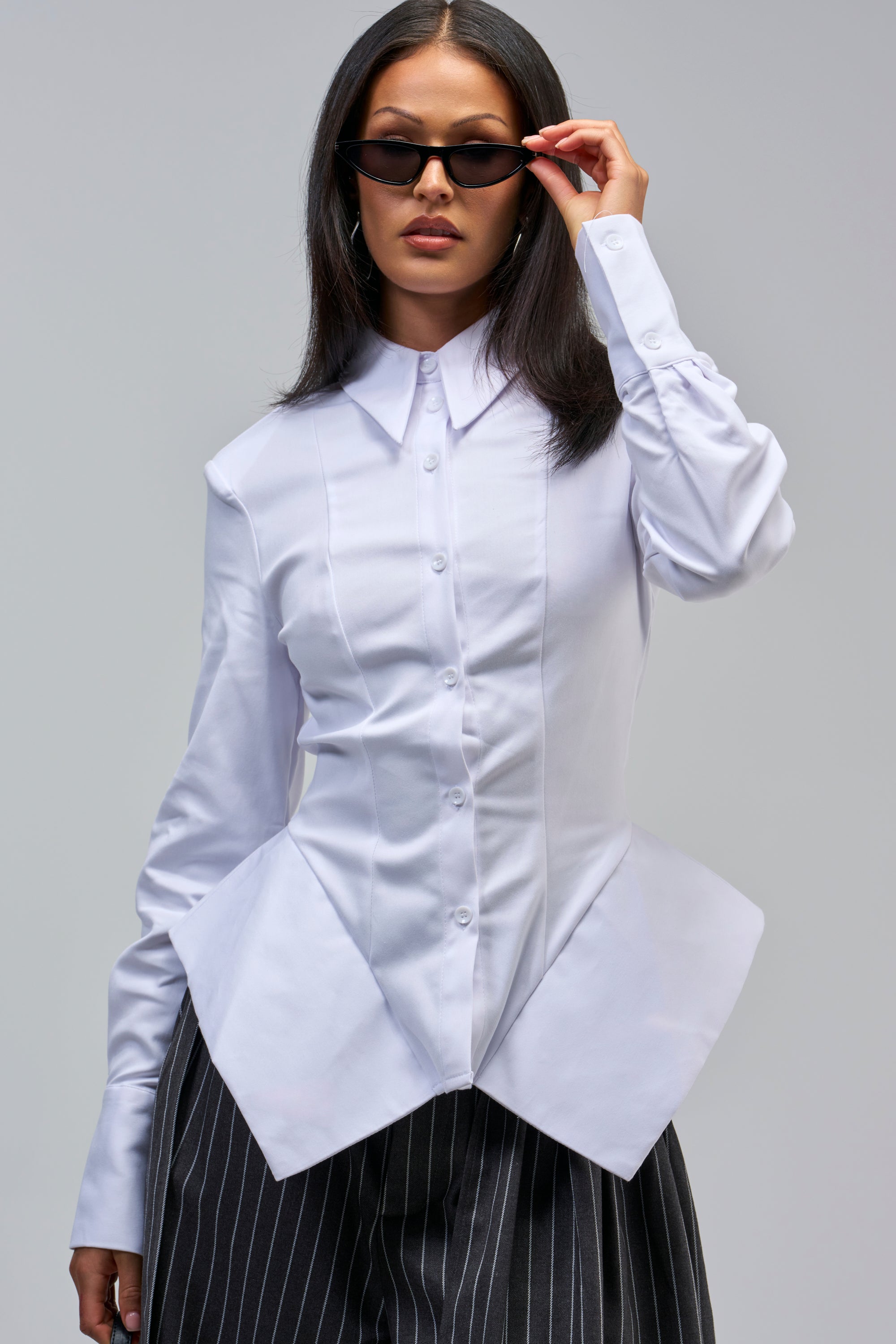 Woman in the OFFICE HOTTIE POPLIN BUTTON DOWN, featuring an angular peplum design, pairs it with pinstripe pants and black sunglasses, posing confidently against a plain gray background.