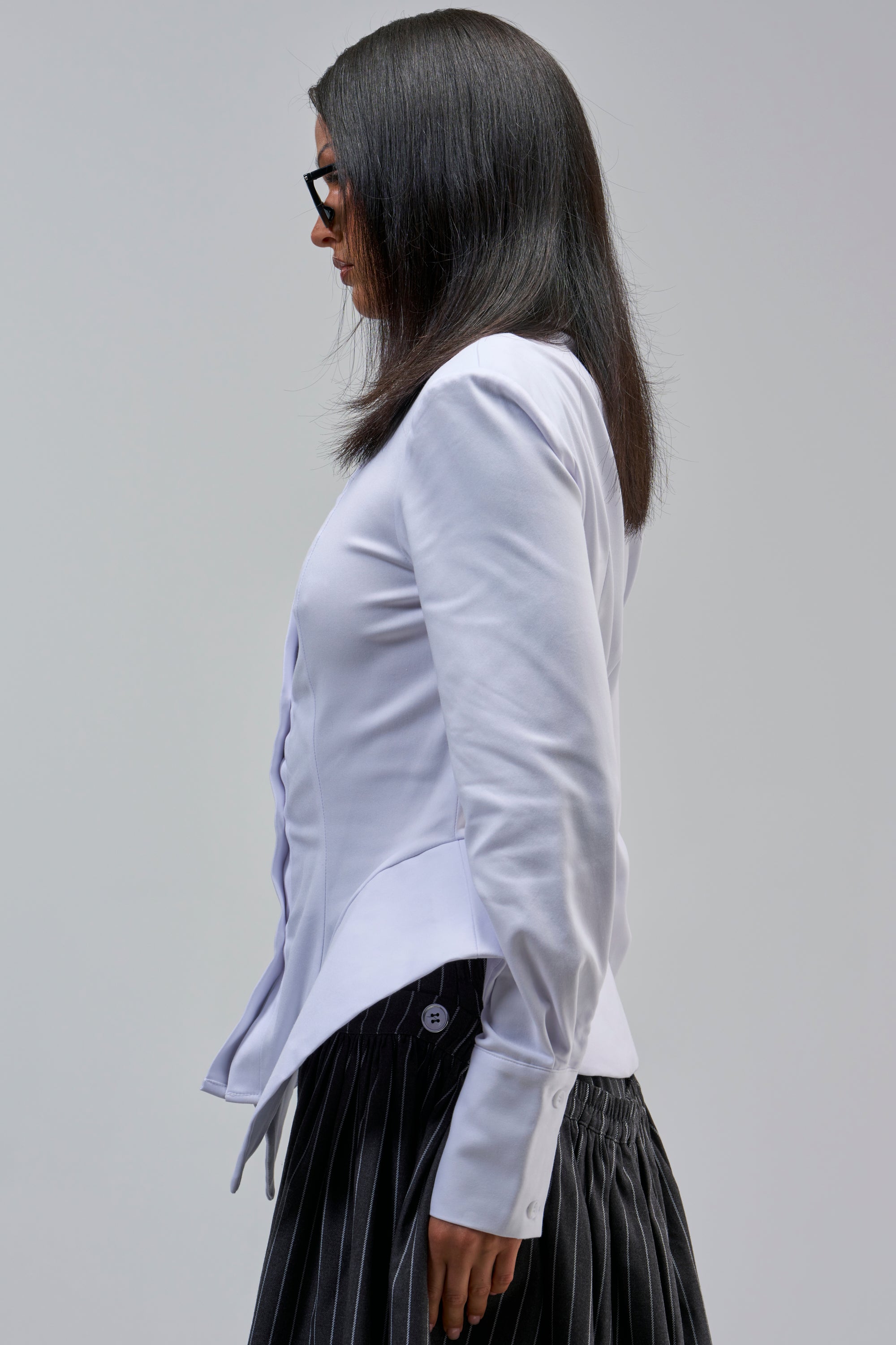 A person with straight dark hair and glasses, wearing the OFFICE HOTTIE POPLIN BUTTON DOWN blouse and a pleated dark skirt, stands in profile against a plain light gray background.
