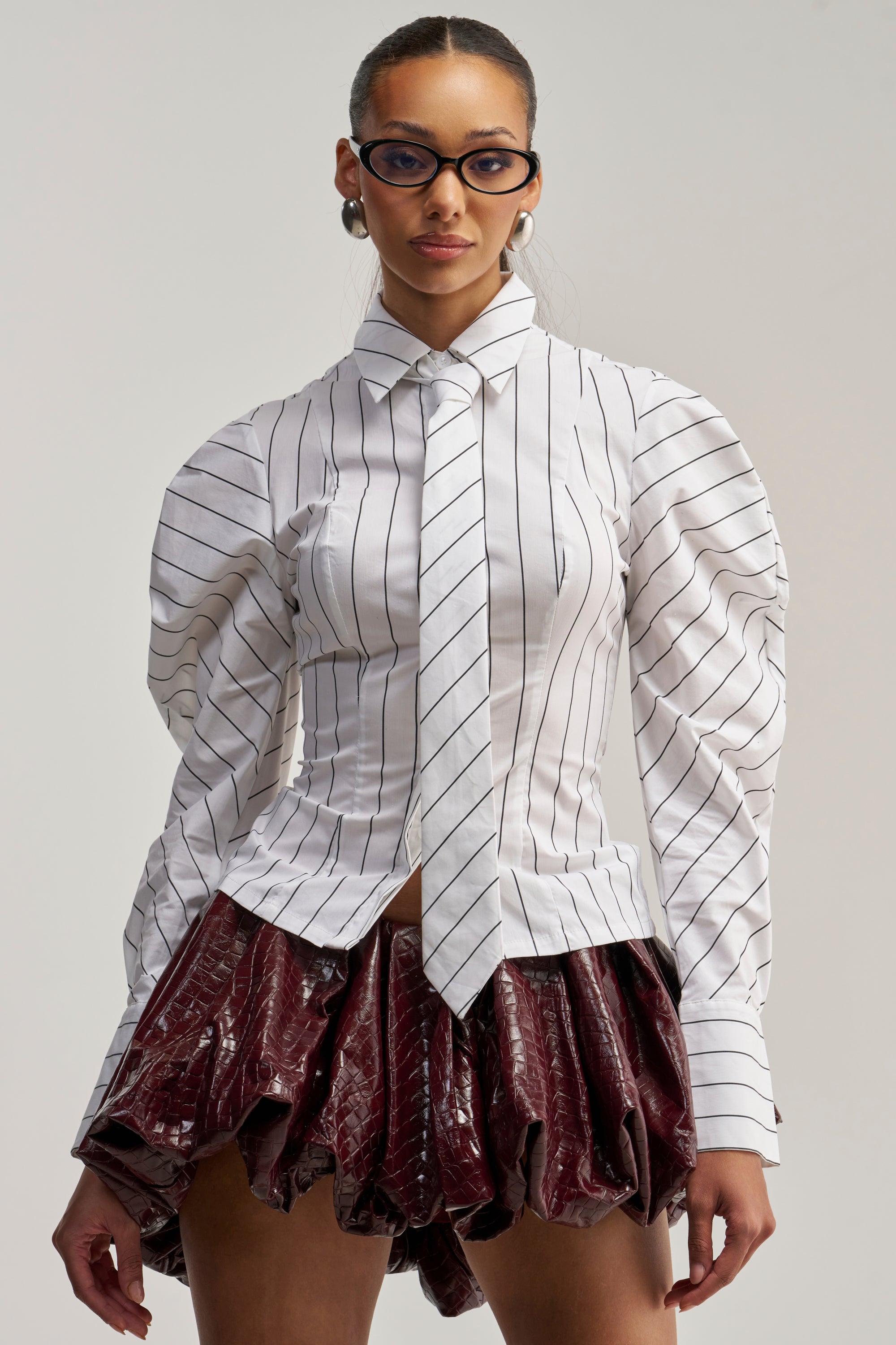 Wearing the “Class Is In Session Poplin Button Down,” a woman styles this crisp white pinstripe shirt with a tie as a sophisticated statement piece. The design features puffed sleeves and is paired with metallic earrings, blue glasses, and a shiny burgundy textured skirt. Standing against a plain background and looking forward, she presents an elegant interpretation of modern workwear.
