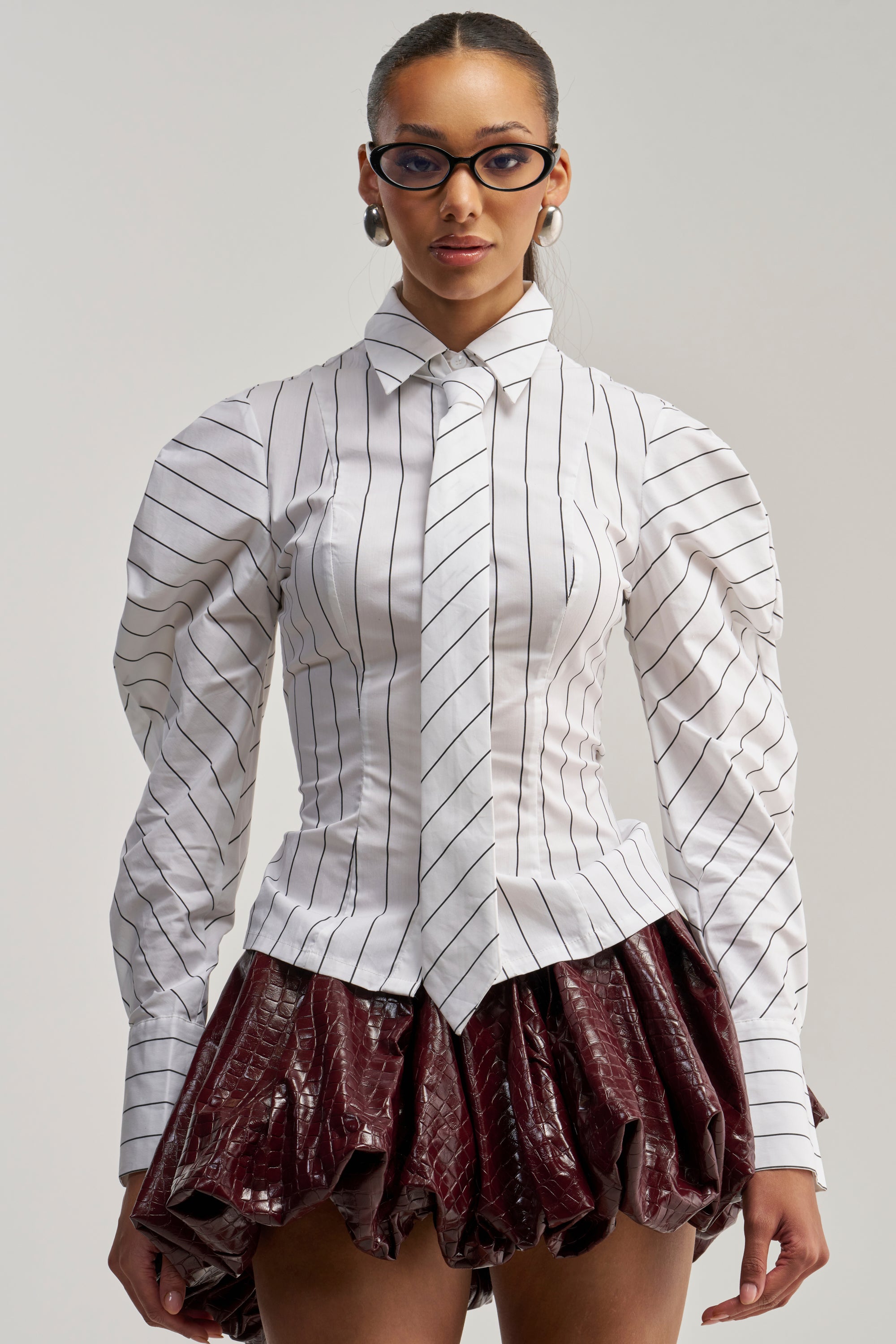 A woman wearing glasses and the CLASS IS IN SESSION POPLIN BUTTON DOWN with puffed sleeves stands against a plain light background, styled with a shiny burgundy skirt.