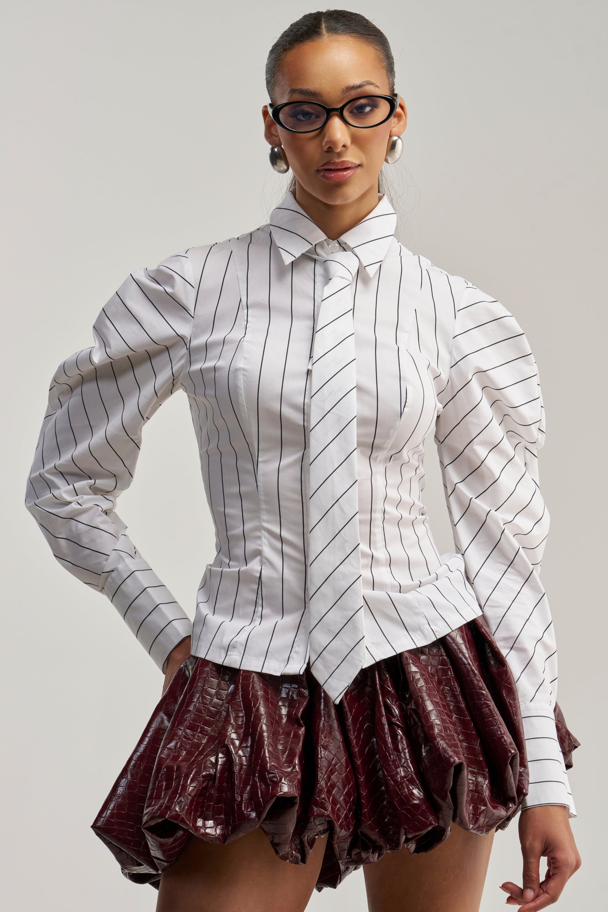 A woman exudes office siren style in the CLASS IS IN SESSION POPLIN BUTTON DOWN, a crisp white pinstripe shirt with puffy sleeves and a detachable tie detail, paired effortlessly with a shiny burgundy textured skirt as she poses confidently against a plain background.