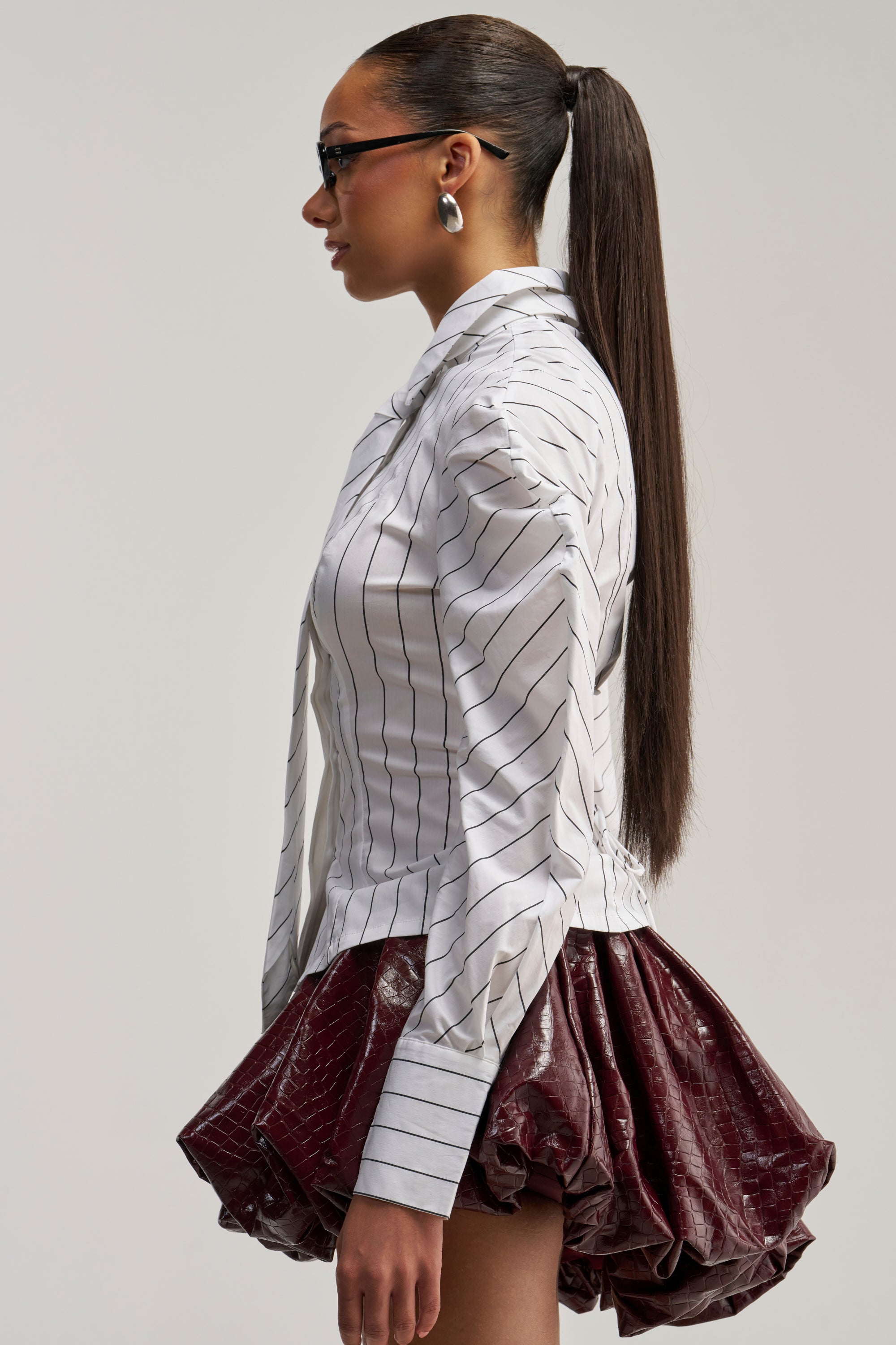 A woman in profile wears glasses and a sleek, long ponytail. She’s styled in the CLASS IS IN SESSION POPLIN BUTTON DOWN, fashioned as a detachable tie blouse, and pairs it with a shiny maroon bubble skirt. Statement earrings emphasize the bold confidence of office siren style against a plain, light-colored background.