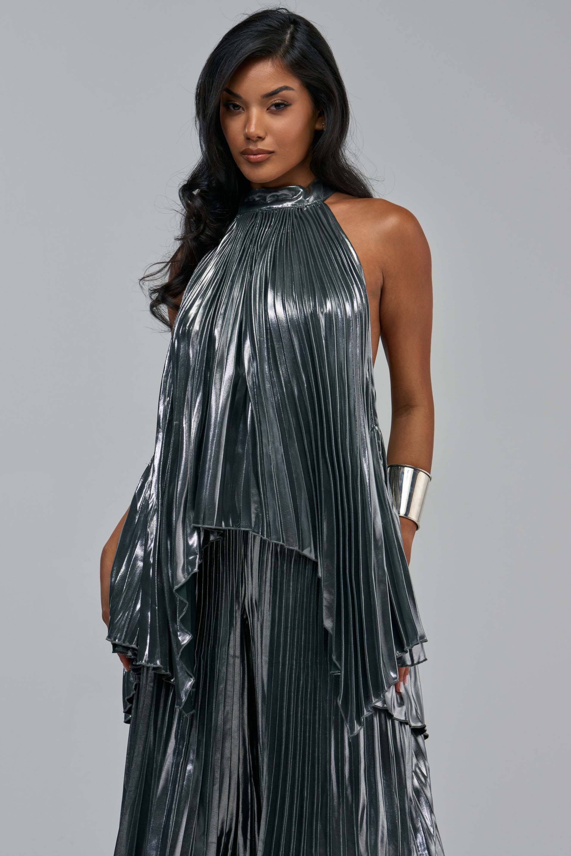 A woman wears the metallic silver ALL EYES ON ME PLEATED TOP, featuring a high halter neckline and a wide silver cuff bracelet.