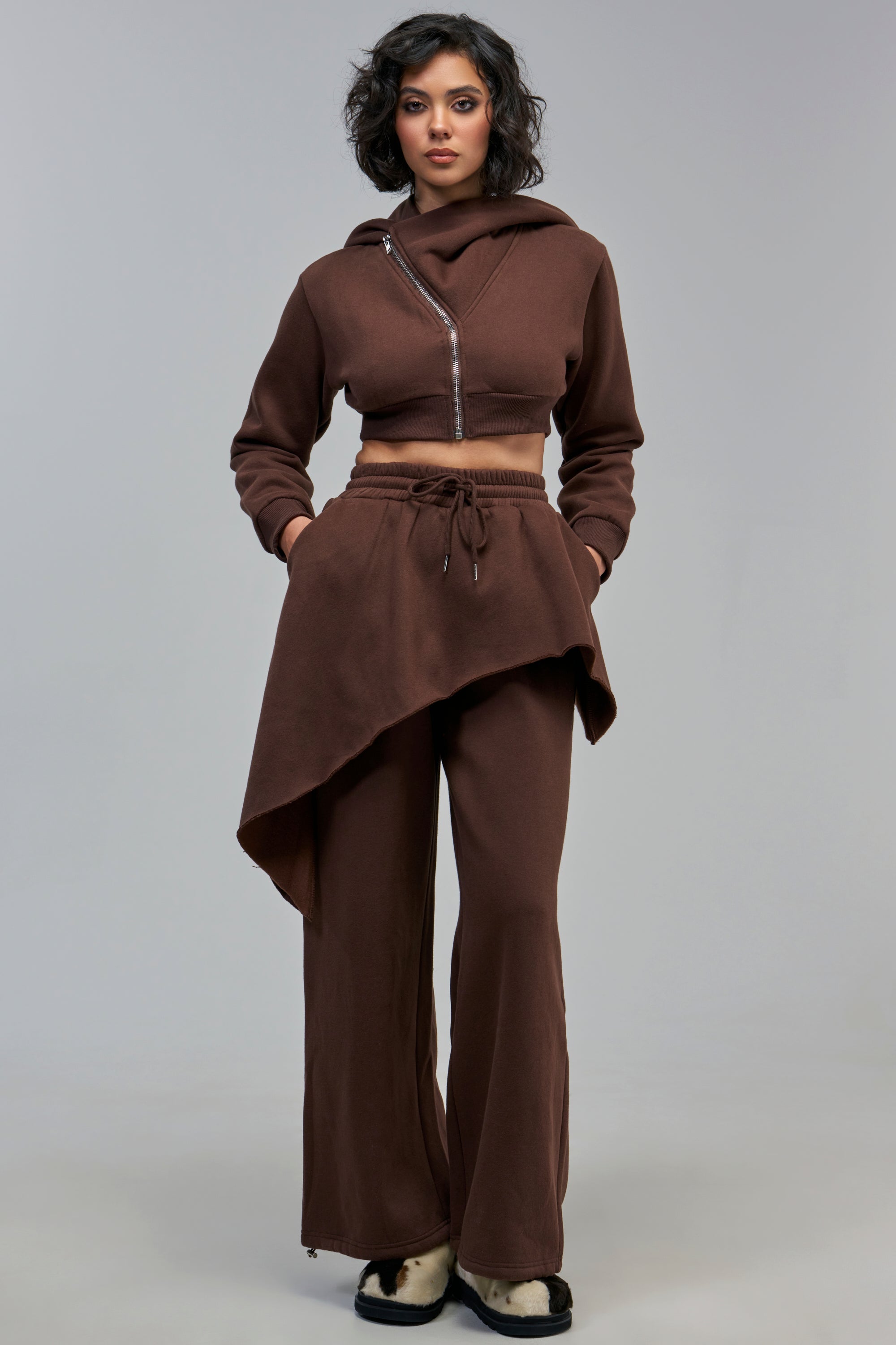 A person wearing a cropped, asymmetrical brown hoodie and matching MISS INDEPENDENT SWEAT PANT with a high-low skirt overlay in a layered streetwear style.