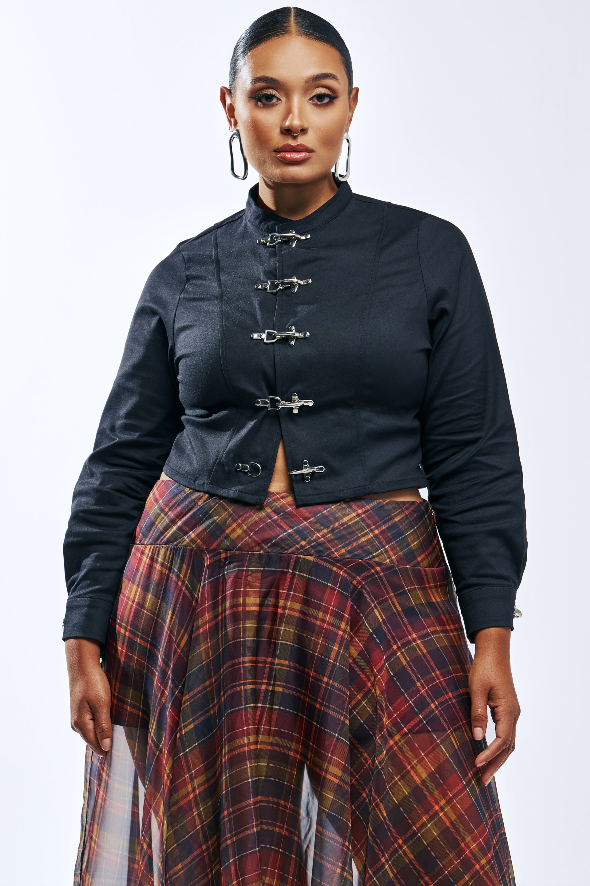 A person wears a black long-sleeved cropped UNBOTHERED BLOUSE with silver toggle closures over a red and yellow plaid skirt.