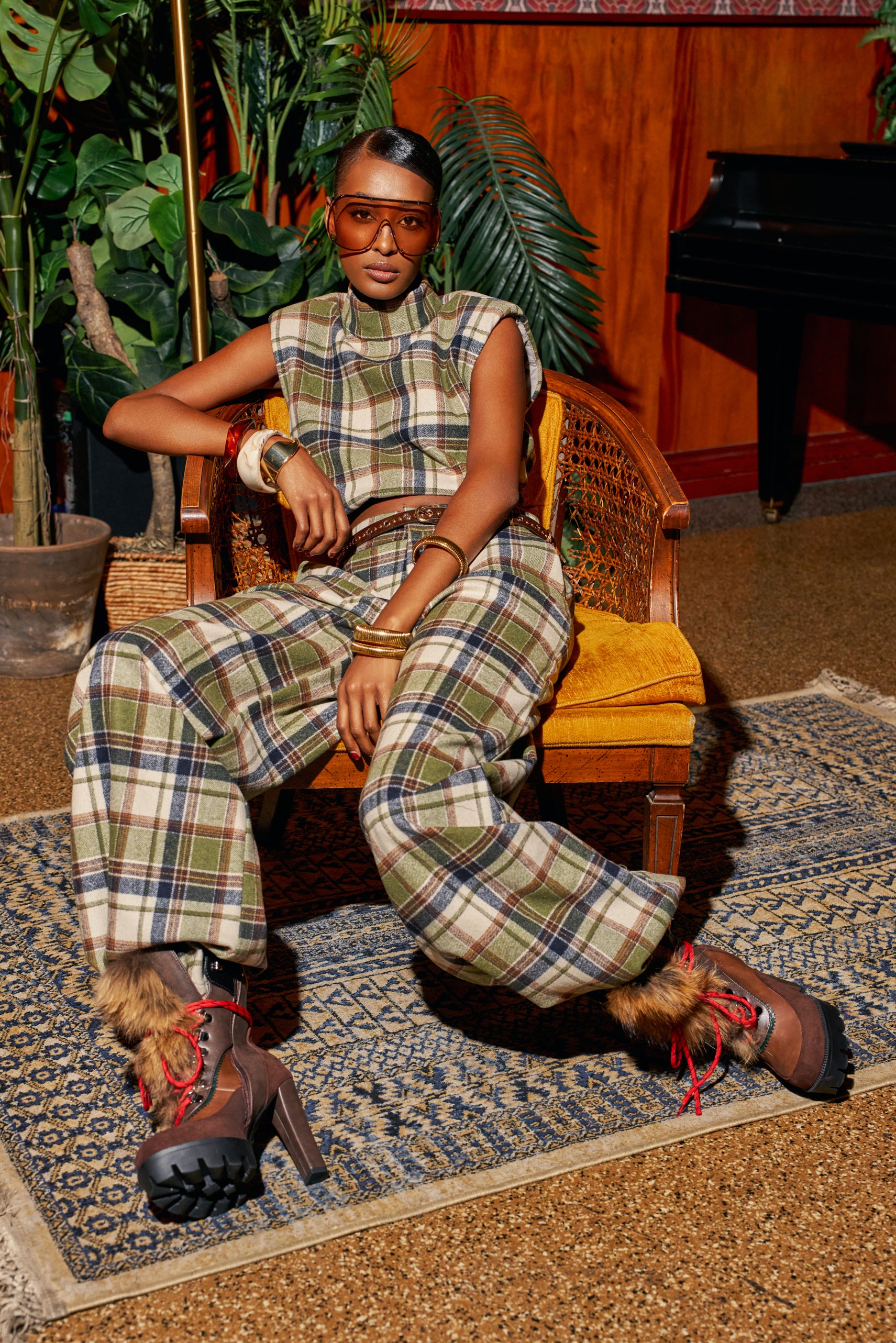 A person wearing a sleeveless plaid top and matching olive green LUCKY ONE PLAID PANT wide-leg trousers, seated in a wooden chair, wearing tinted oversized glasses and heeled hiking boots with faux fur trim.