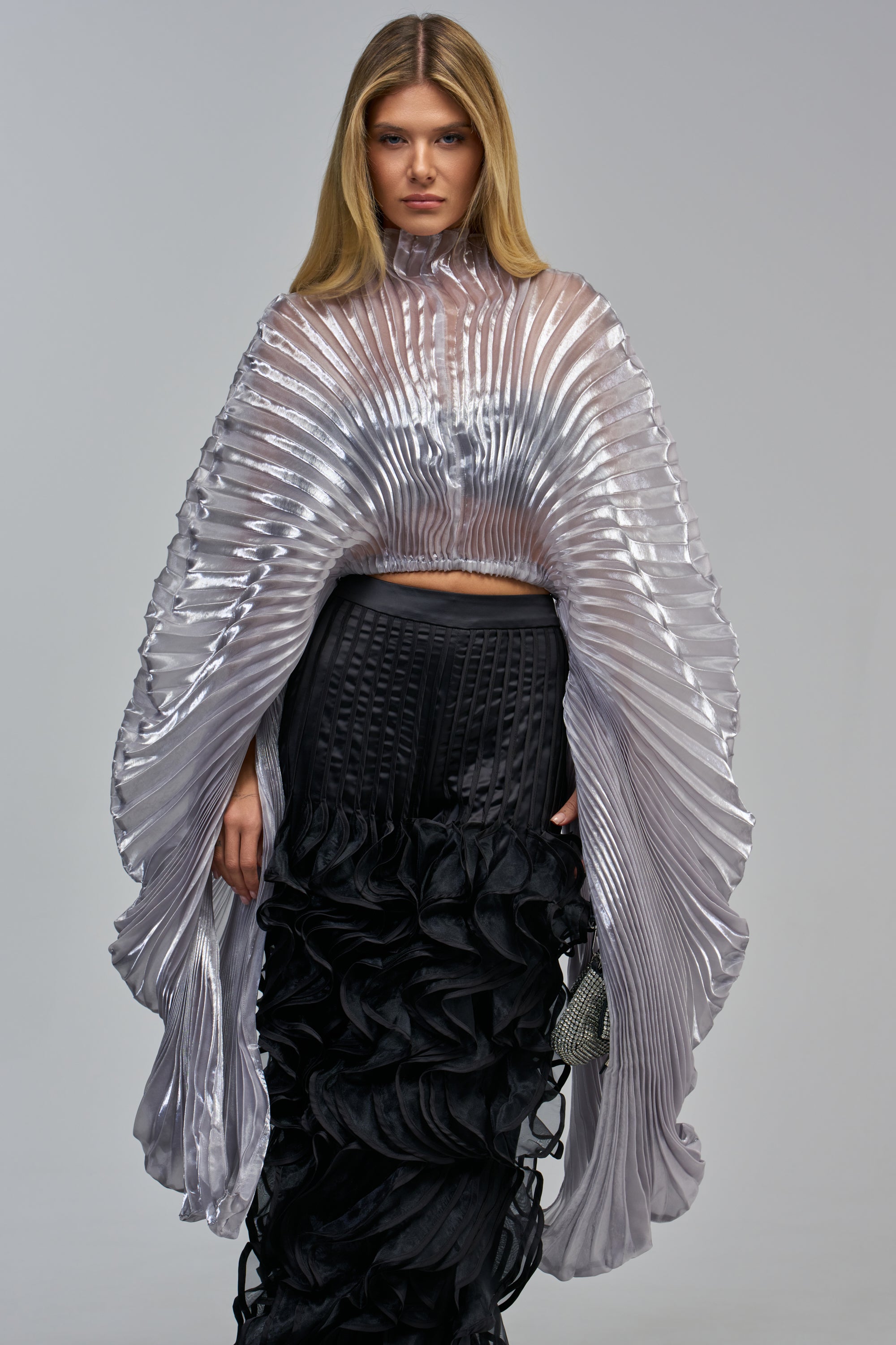 A model wears the PASS THE BUBBLY PLEATED TOP IN SILVER, featuring a high-neck, metallic silver pleated fan design with dramatic, wide bell sleeves, paired with a black tiered ruffle skirt for luxe evening styling.